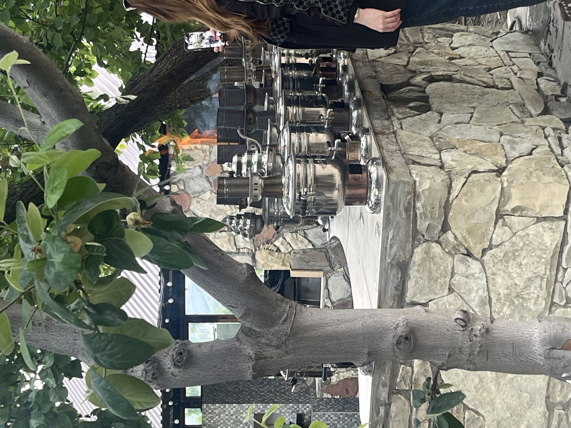 The image shows a display of shiny, metal samovars, possibly for sale or exhibition. A person is taking a photo of the display with their smartphone. The background consists of stonework, suggesting an outdoor setting. A tree with green leaves and branches frames the left side of the image. The image is shot from a ground-level perspective, with a slight upward angle. The location is identified as the city of Miatli, Russia.