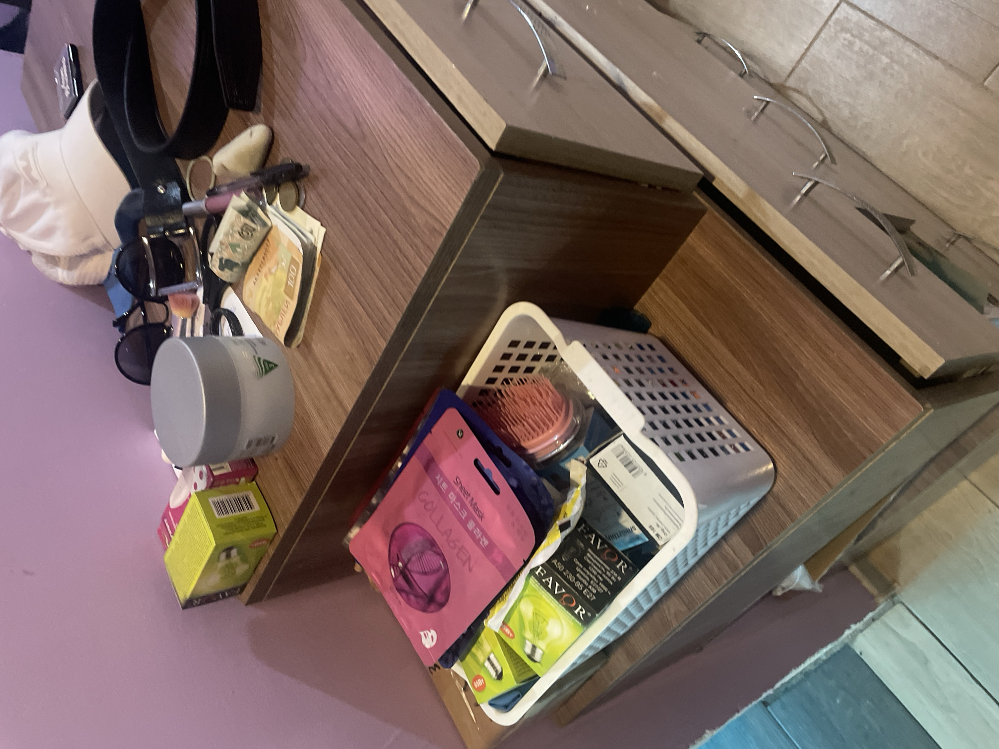 This is an indoor scene, likely in Olginka, Russia, featuring items on and under a wooden cabinet with drawer pulls.
On the top surface, there's a mix of personal items: a white cap, a black belt, sunglasses, cosmetics, and Belarusian currency (РУБЛЕЙ).
Under the cabinet, there is a white plastic basket containing a pink sheet mask ("COLLAGEN"), a hairbrush, and packaging for "FAVOR" light bulbs. A round container sits on top of a box of the same light bulbs.