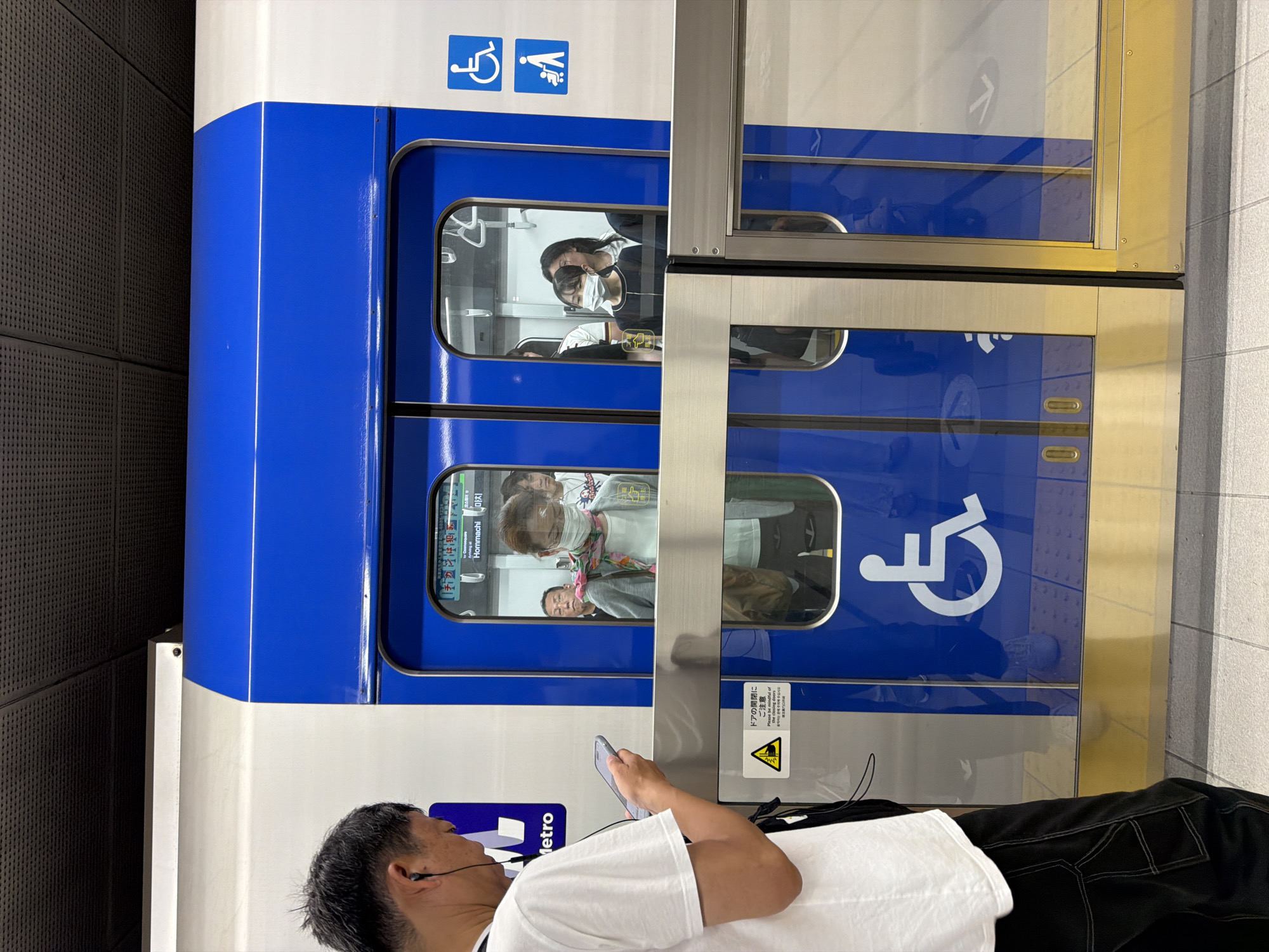 The image captures a bustling scene at a train station platform, with a blue and white train docked. In the foreground, a man with short dark hair, wearing a white t-shirt and dark pants, is seen from behind, engrossed in his smartphone. He has wired earphones plugged in, suggesting he might be listening to audio.
The train car features a distinctive blue stripe along its windows and a white upper body. Through its windows, several passengers are visible inside. One woman with long dark hair, wearing a dark top and a white face mask, looks out towards the viewer. Another person, also wearing a mask and a colorful scarf, is partially visible.
Above the train windows, international accessibility symbols are displayed: a wheelchair user icon and a priority seating symbol. The word "Metro" is partially visible on the train's side.
The platform is separated from the train by transparent safety barriers. On one of these barriers, a prominent yellow triangular warning sign is visible. It features an exclamation mark and a pictogram of a person caught in a door, with text in both Japanese and English. The Japanese text reads "ドアの開閉にご注意" (Doa no kaihei ni go-chūi), meaning "Please be careful of the door opening/closing," followed by the English translation: "Please be mindful of door."
A digital display inside the train, visible through one of the windows, indicates "Hommachi" and its Korean transliteration "마치" (Machi), suggesting the train's location or destination is likely a station called Hommachi, possibly in Japan. The background features a dark, perforated wall. The overall scene depicts a typical moment in an urban transit system.