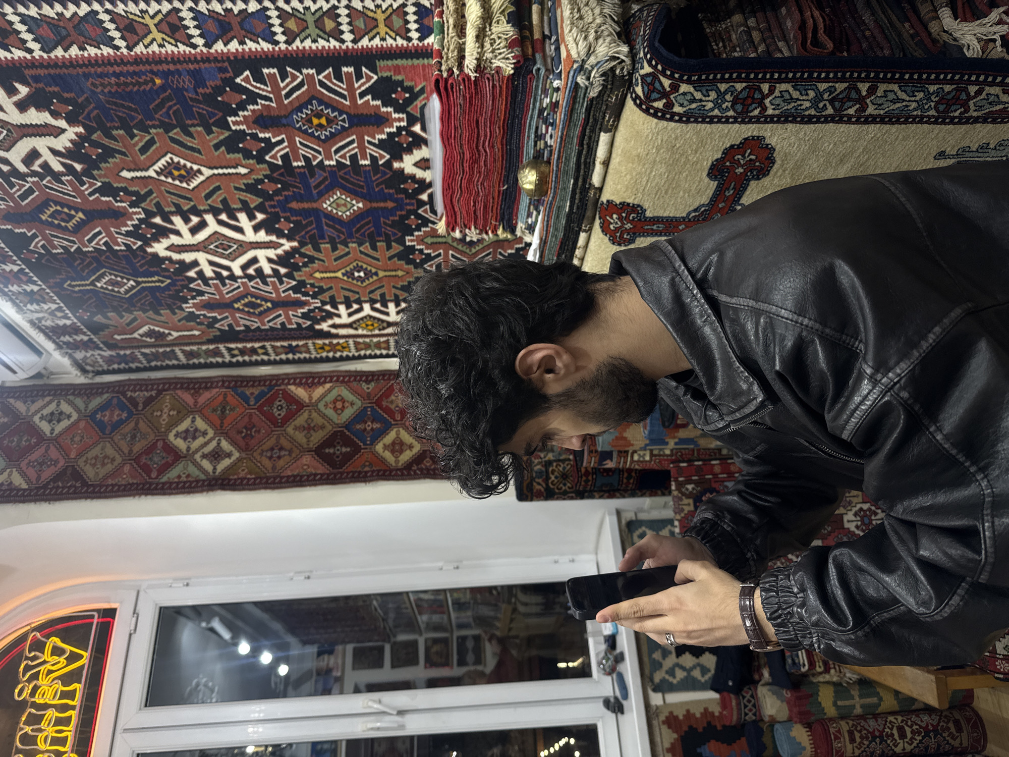 A person wearing a black leather jacket is looking at a mobile phone in a shop in Yerevan, Armenia. The shop is filled with carpets hanging on the wall and stacked on the floor. The person is standing in front of a window. A neon sign can be seen in the window. The person has short dark curly hair, a beard, and is wearing a ring on their finger. A bracelet is visible on the person's wrist.