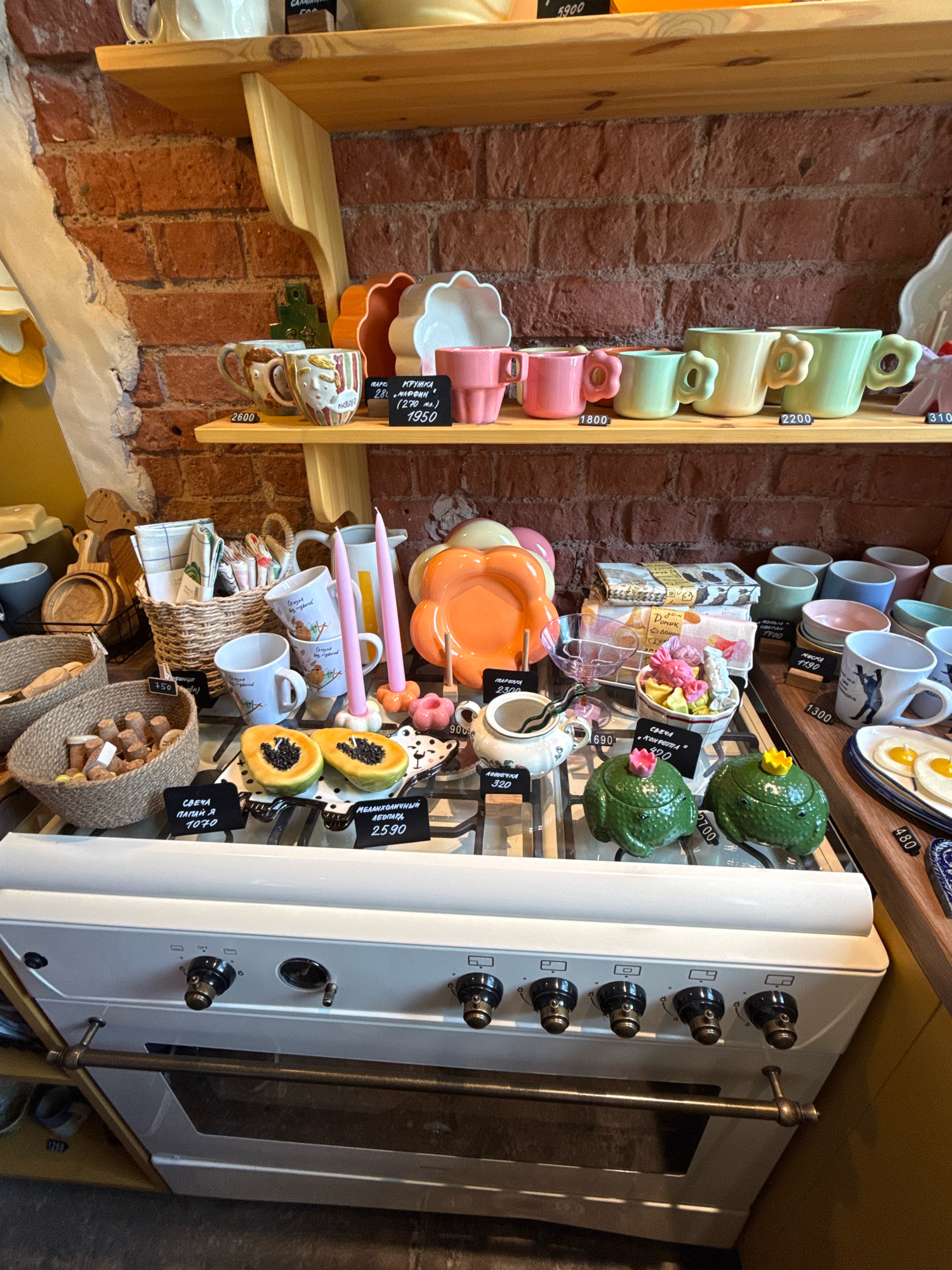 The image shows a retail display of ceramics and home goods on shelves above a white stove. The merchandise includes mugs with intricate designs, pastel-colored cups, decorative bowls, candles, and ceramic figurines shaped like frogs and a pumpkin. Some items have price tags. The background is a brick wall. A sign in the background indicates the location is "Design District Kaliningrad," a concept store in Kaliningrad, Russia, located at Ulitsa Lenina, 14a. There are no people visible in the image.