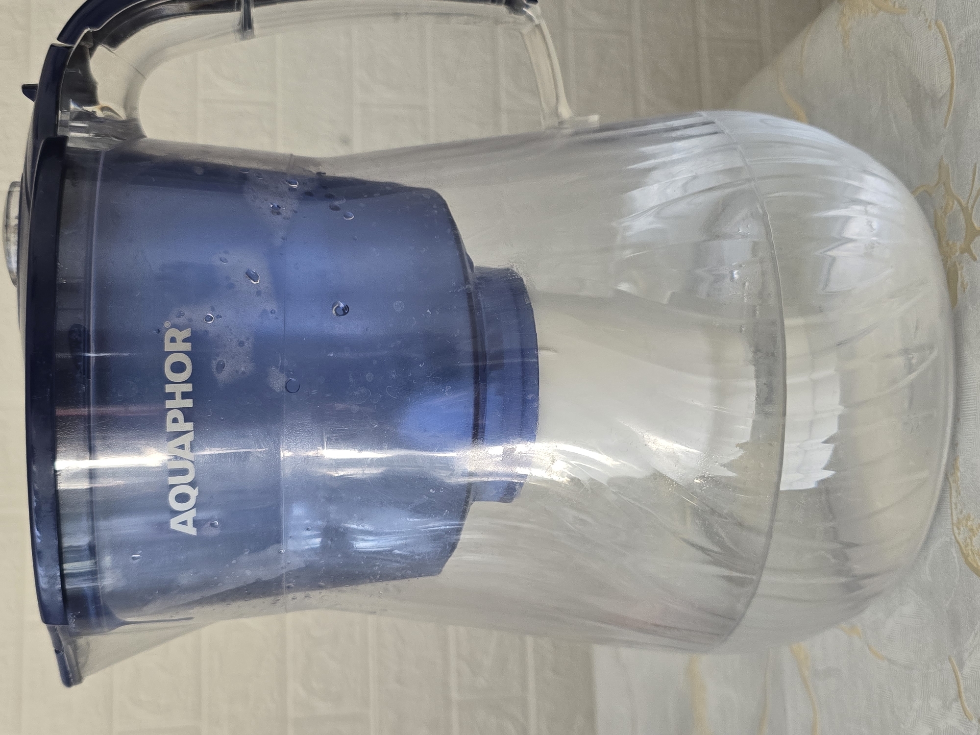 A full shot from a slightly elevated perspective shows a clear plastic Aquaphor brand water filter pitcher filled with water. The pitcher is dark blue at the top, with the brand name "AQUAPHOR" prominently displayed in white lettering. A blue filter housing is submerged in the pitcher, with a white filter cartridge visible inside. The pitcher is placed on a patterned tablecloth in front of a white brick wall. The lighting is even, suggesting an indoor setting. The image does not depict any people or specific environmental features that would indicate a location within the city of Qaratau, Kazakhstan.