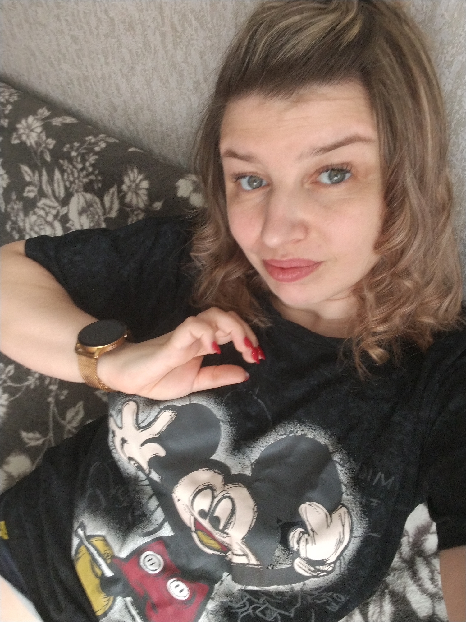 A woman is lying on a couch with a gray floral pattern, wearing a black Mickey Mouse t-shirt and a gold watch. She is looking at the camera with a neutral expression. Her hair is blonde and curly, and she has red nail polish on. The location is unclear, but no specific details suggest the city of Yaroslavl, Russia.