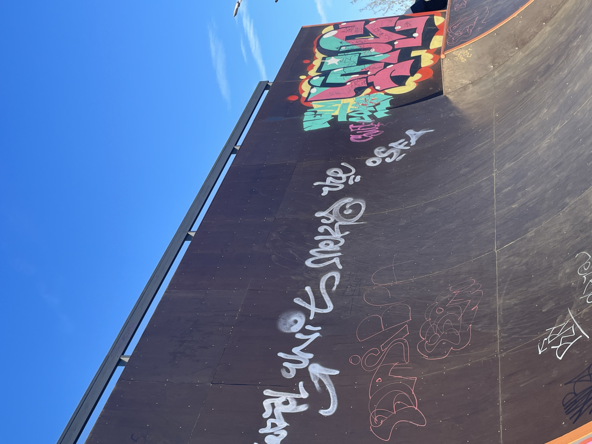 An outdoor skateboard ramp, likely a half-pipe or quarter-pipe, is depicted under a bright, clear blue sky in Izhevsk, Russia. The ramp surface is dark, possibly made of wood or composite material, and is heavily adorned with graffiti.
In the upper-right section, a large, colorful graffiti piece dominates with a vibrant mix of pink, green, yellow, and black outlines. Though highly stylized, words like "REAL" and possibly "LIME" or similar shapes are discernible within the complex design. Below this, smaller purple text is visible.
Further down the dark surface of the ramp, white spray paint graffiti stands out. One clearly legible tag reads "O'SEA" with "TRUE" nearby. Below these, a larger, more intricate white tag is painted. Towards the lower-right curve of the ramp, additional graffiti is present, primarily in red outlines.
The image is captured on a sunny day, indicated by the brilliant blue sky, which shows only a few wispy white clouds and a small bird flying in the distance. A metal railing or coping runs along the top edge of the ramp on the left side, contrasting with the dark surface. The ramp's structure includes a curved section on the right, typical of skatepark features. No people are visible, but the setting suggests a vibrant urban sports environment.