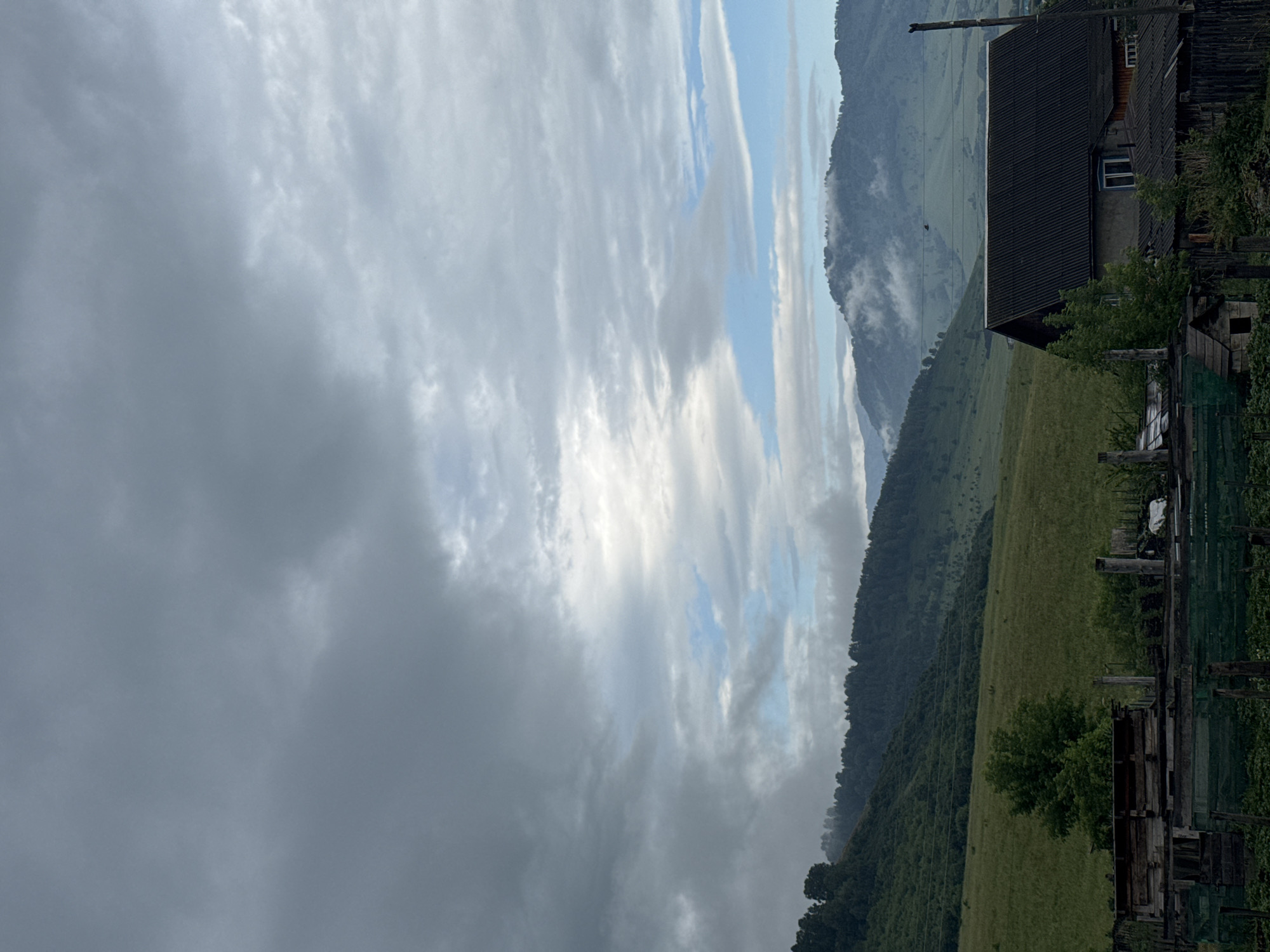This is a vertical, outdoor, eye-level shot of a cloudy sky above a rural hillside in Soloneshnoye, Russia. The sky is dominated by large, dramatic clouds with patches of blue sky peeking through. Sunlight is breaking through the clouds in places, creating bright highlights. The clouds are a mix of dark gray and lighter white, suggesting a potentially changing weather pattern.
The scene below the sky features a steep, tree-covered mountain slope that transitions into a grassy field. There are visible power lines stretching across the hillside. In the foreground, on the right side of the image, is a portion of a rustic wooden house with a dark, corrugated roof. A smaller wooden structure, possibly a shed or animal enclosure, is also visible. A wooden fence encloses part of the property.
The time of day appears to be either early morning or late afternoon, given the quality of light filtering through the clouds and the soft shadows. The overall mood is serene and natural, with a sense of isolation and natural beauty. No people are visible in the image. No text is discernible.