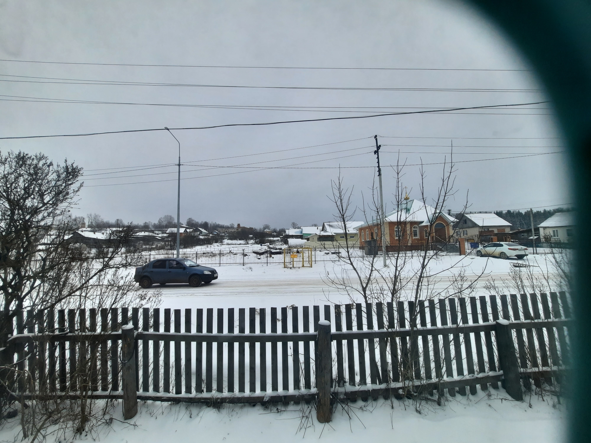 The image shows a winter scene in what appears to be Krasnogorskoye, Russia, based on the architecture of the buildings. A dark blue sedan is driving on a snow-covered road. A weathered wooden fence is in the foreground, partly obscured by bare bushes. Beyond the fence are houses, a light post, power lines, and a church-like building with a white dome. The sky is overcast and gray. Snow covers the ground. Another white car is parked nearby the church.