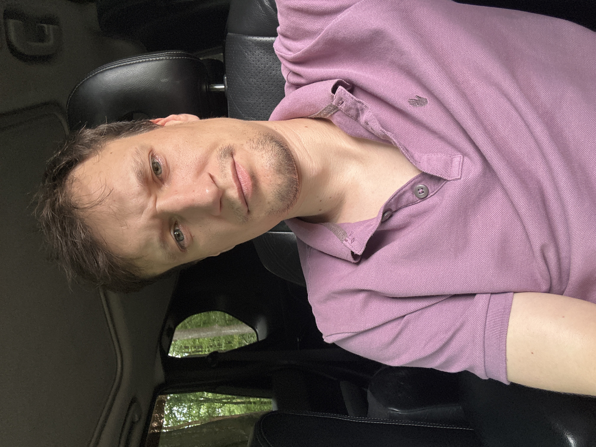 A medium shot selfie is taken from inside a car. The subject is a fair-skinned man with short brown hair and a light beard. He is wearing a light purple polo shirt with a buttoned collar. The man is seated in the driver's seat of a car, with the interior visible, including black leather seats and the roof. Through the car windows, a wooded area with green trees is visible, suggesting an outdoor setting, possibly in or near Shatava, Ukraine, given the context. The man's facial expression is a slight grimace. The lighting is natural and appears to be daytime.