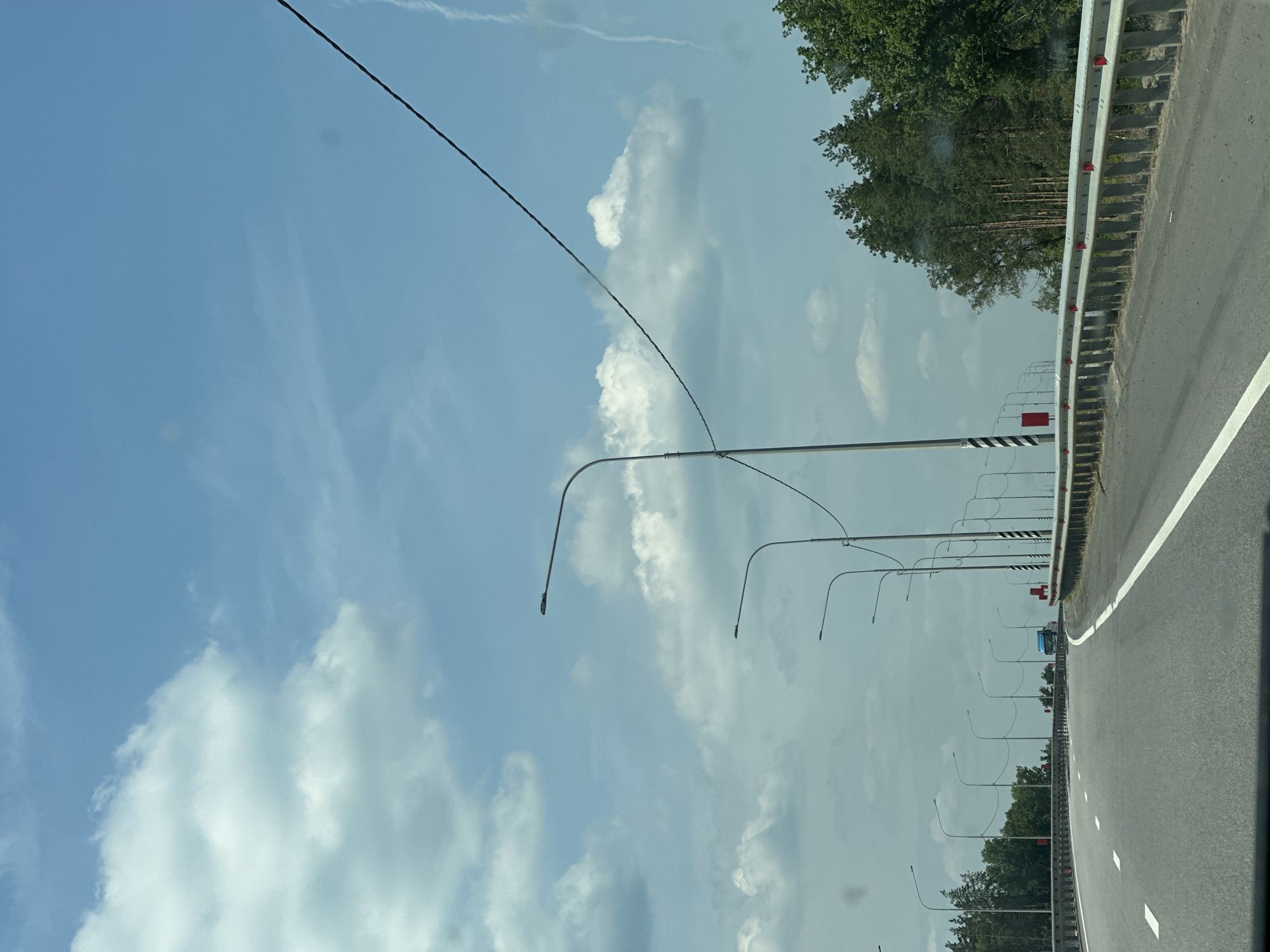 The image captures a forward view from a moving vehicle on a highway, likely a bridge or elevated roadway, given the guardrails and the perspective. The sky is blue with scattered white clouds. A series of streetlights are visible along the right side of the road, extending into the distance. A power line runs diagonally across the upper portion of the frame. On the right, a dense line of coniferous trees forms the edge of the landscape. The highway has clear white lane markings. A blue truck is visible in the distance on the highway. The location is identified as Rasskazovo, Russia.