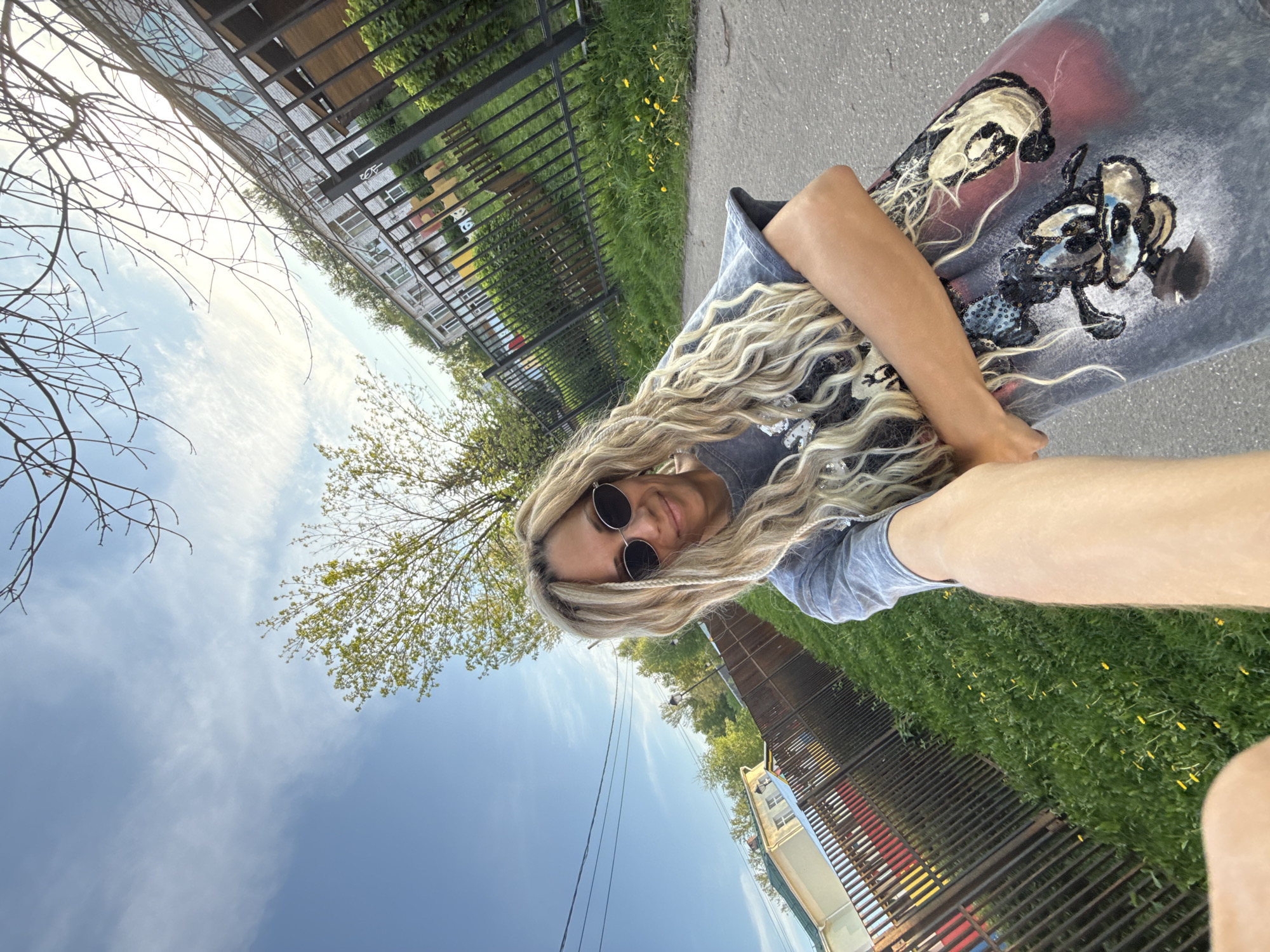 A young woman stands along a fence, wearing sunglasses and a t-shirt with a graphic design on it. Her long, blonde hair hangs down. She's near a grassy area and what appears to be a sidewalk or paved area. In the background are buildings and trees, suggesting an urban setting. Based on the request for a location within Rusanovo, Russia, this could be a park or residential area in that city.