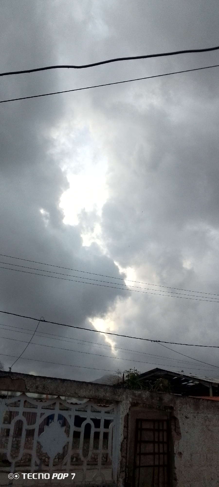 The image displays an overcast sky above a residential scene in Altagracia, Venezuela. The sky is largely covered by heavy, dark grey clouds, which dominate the upper two-thirds of the frame. A significant bright white area, indicative of sunlight breaking through or reflected, stands out prominently in the upper-middle and central portions of the sky.
Multiple black utility wires, likely power or telephone lines, stretch across the sky horizontally, with some wires running parallel to each other. In the lower section of the image, the top portion of a concrete wall is visible. The left side features a white section with a decorative, patterned cutout design. To the right, the wall appears more weathered, with a darker, rougher texture and a section containing a dark, metal-barred opening, possibly a window or gate.
Above the wall, faint outlines of hills or mountains can be seen in the distance, partially obscured by the clouds. Sparse green foliage and parts of a dark roof structure are also visible peeking over the wall. Several tiny, dark specks, possibly birds or insects, are scattered against the brighter areas of the sky. No people are visible, and no specific activity is taking place. The heavy cloud cover suggests an overcast day, potentially signaling rain or an impending storm. A watermark reading "TECNO POP 7" is present in the bottom-left corner.