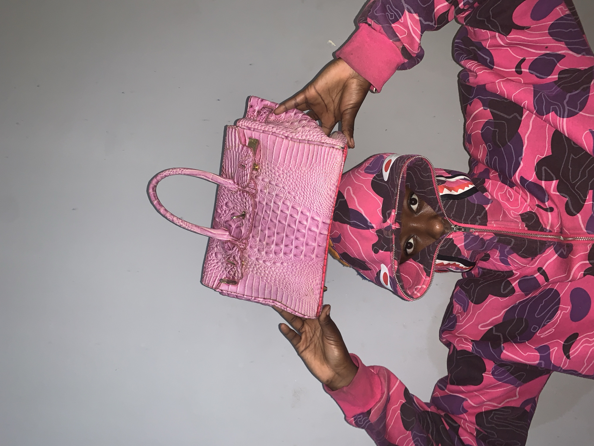 The image shows a person wearing a pink and purple camouflage hoodie with a shark design on the hood. They are holding a pink crocodile-textured handbag. The background is a light gray surface. The location and city are not discernible from the provided image.