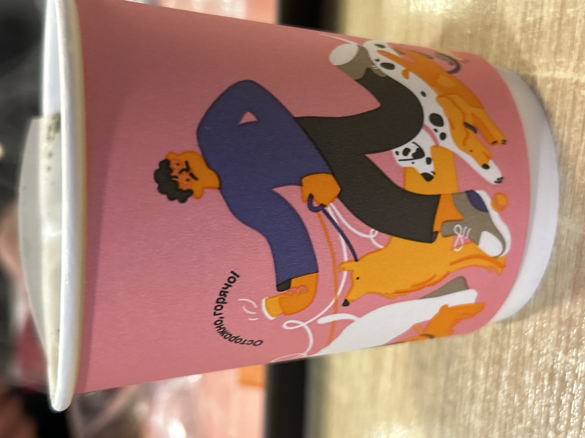 A disposable paper cup, featuring a vibrant pink background, rests on a light-colored surface, possibly wood. The cup is adorned with a dynamic illustration depicting a person, appearing to be a man, attempting to walk a group of energetic dogs. The man is dressed in a blue top, dark trousers, and orange shoes, and his focused expression suggests the effort involved in managing the lively canines.
He holds several leashes, visible as white lines, which extend to multiple dogs running in different directions. The dogs are varied in appearance; one prominent dog is white with black spots, resembling a Dalmatian, while others are predominantly orange or brown, and one is white. The illustration's style is cartoonish and minimalist, emphasizing the playful chaos of dog walking.
The top of the cup features a clean white rim. Curved along the side of the cup is visible text in Russian: "осторожно, горячо!" (ostorozhno, goryacho!), which translates to "caution, hot!" or "careful, hot!", indicating the cup's purpose for hot beverages. The scene captures a common urban activity with an engaging design. This item is from Chaikovsky, Russia.