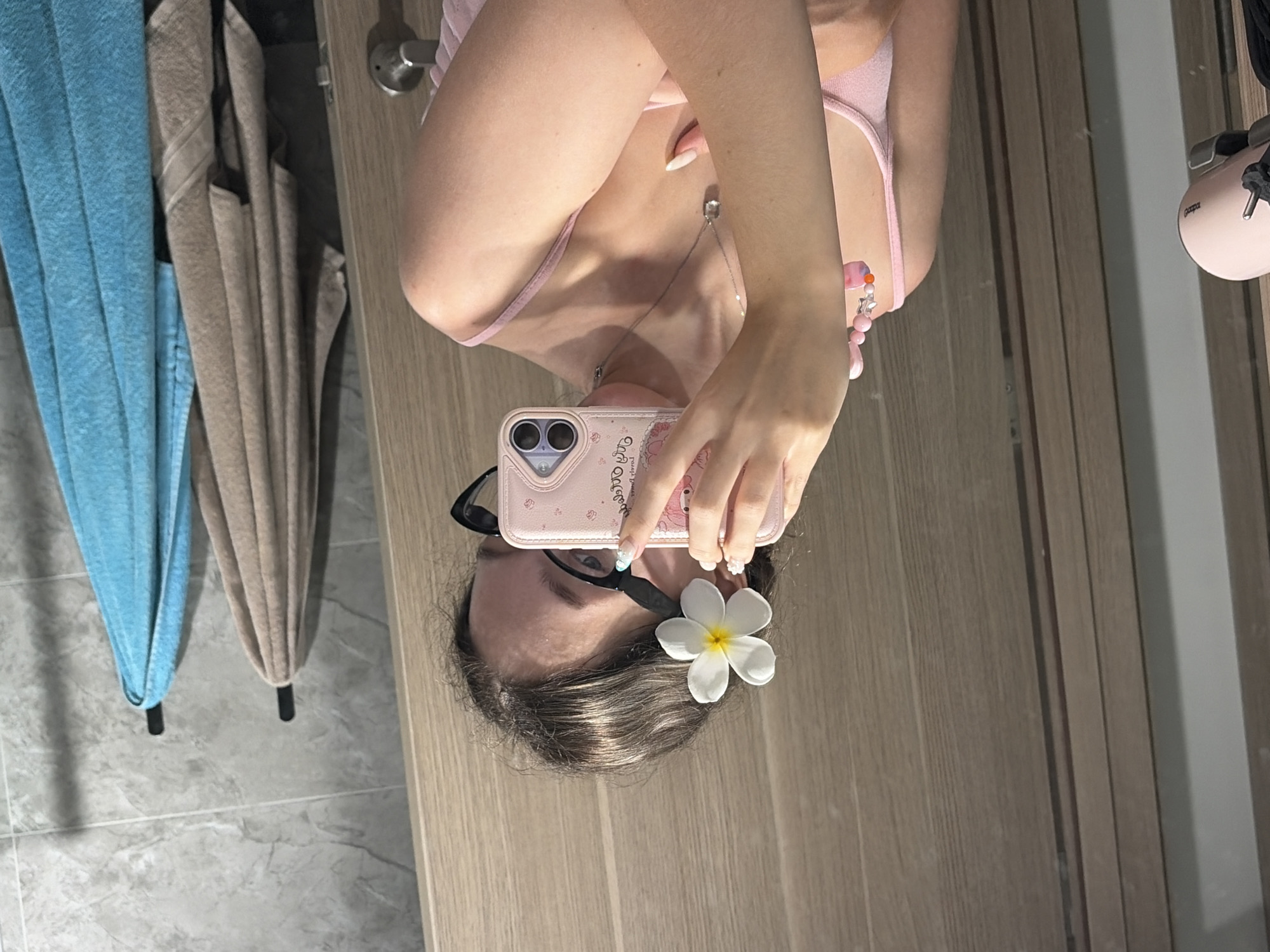 A young woman is taking a mirror selfie in a bathroom. She is wearing a pink tank top and has a white flower tucked behind her ear. She is holding up a pink phone with a cartoon design, obscuring her face. The background shows a wooden door, a tiled wall with two towels hanging on hooks, and a hairdryer attached to a wall. The lighting is bright, suggesting an indoor setting, possibly a hotel bathroom. There is no specific information in the image to suggest the location is Hà Nội, Vietnam.