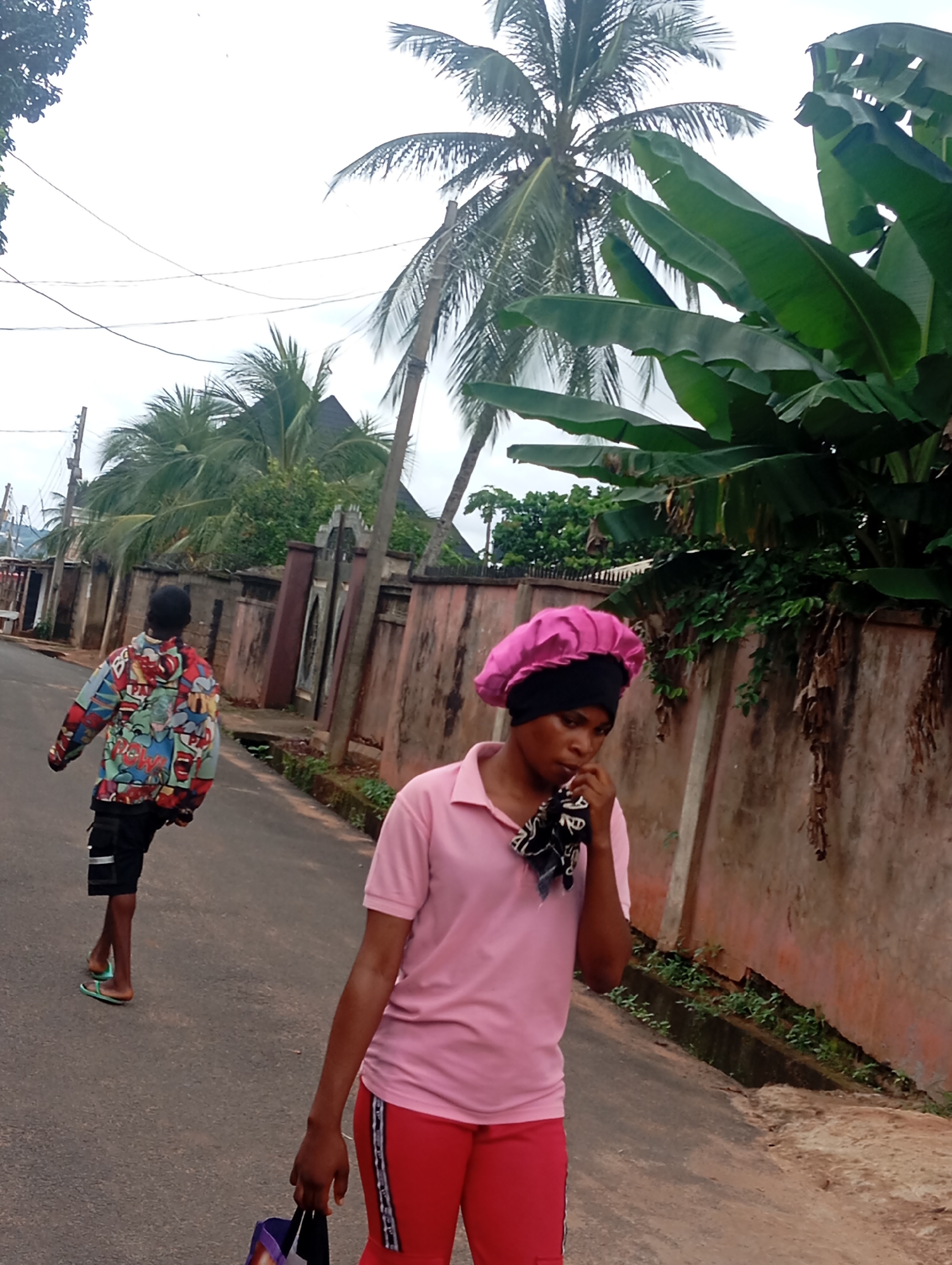 The image depicts a street scene in Nsugbe, Nigeria, with two individuals walking on an asphalt road. In the foreground, a person wearing a pink polo shirt and red pants is walking away from the camera, holding a patterned cloth to their mouth. They have a pink head covering and are carrying a bag in their left hand. Further down the road, another individual is walking away from the camera, facing towards a colorful, graphic-patterned jacket and black shorts. This person is wearing flip-flops. The environment includes a weathered, reddish-pink wall lining the right side of the road, with various green foliage and banana leaves visible overhead. A tall palm tree is also prominent in the background, along with power lines and some structures that appear to be houses or buildings. The sky is overcast.