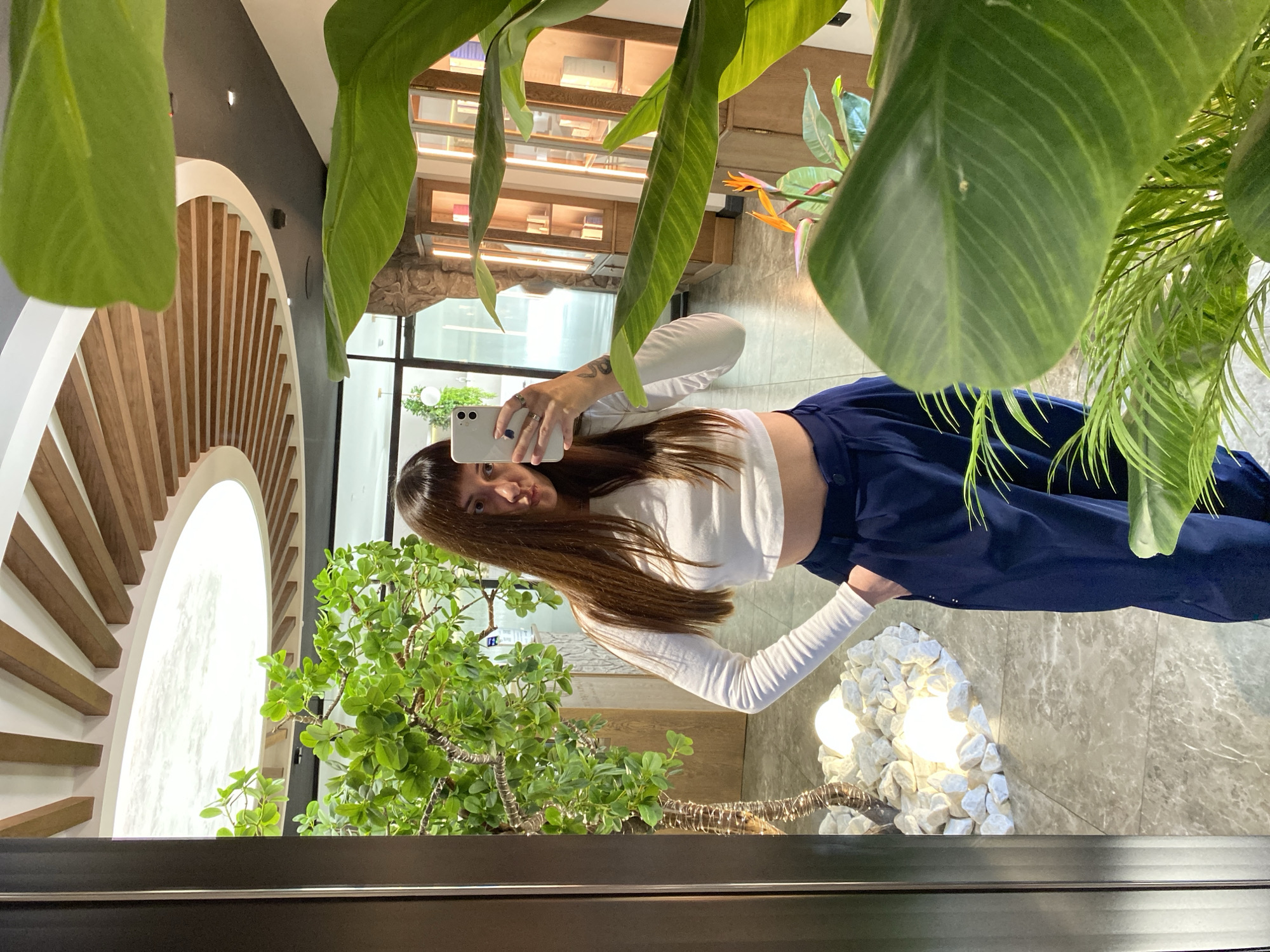 A full-body mirror selfie is taken by a young woman with long, dark brown hair and bangs. She is wearing a white long-sleeved crop top and dark blue wide-leg pants. Her left hand is in her pocket, and her right hand is holding a white smartphone, capturing the reflection. The environment appears to be an indoor space with polished gray tiled flooring. To the left, a decorative tree with vibrant green leaves is illuminated by two round white lights nestled among white stones. The background shows a retail or display area with wooden cabinets and shelves showcasing various products. Large green leaves of a tropical plant partially obscure the right side of the frame. The ceiling features a unique design with concentric wooden beams radiating from a central circular light fixture. The setting is consistent with a modern retail establishment or a well-decorated public space. There is no indication or possibility of determining the location as Votkinsk, Russia based on the visual information provided.