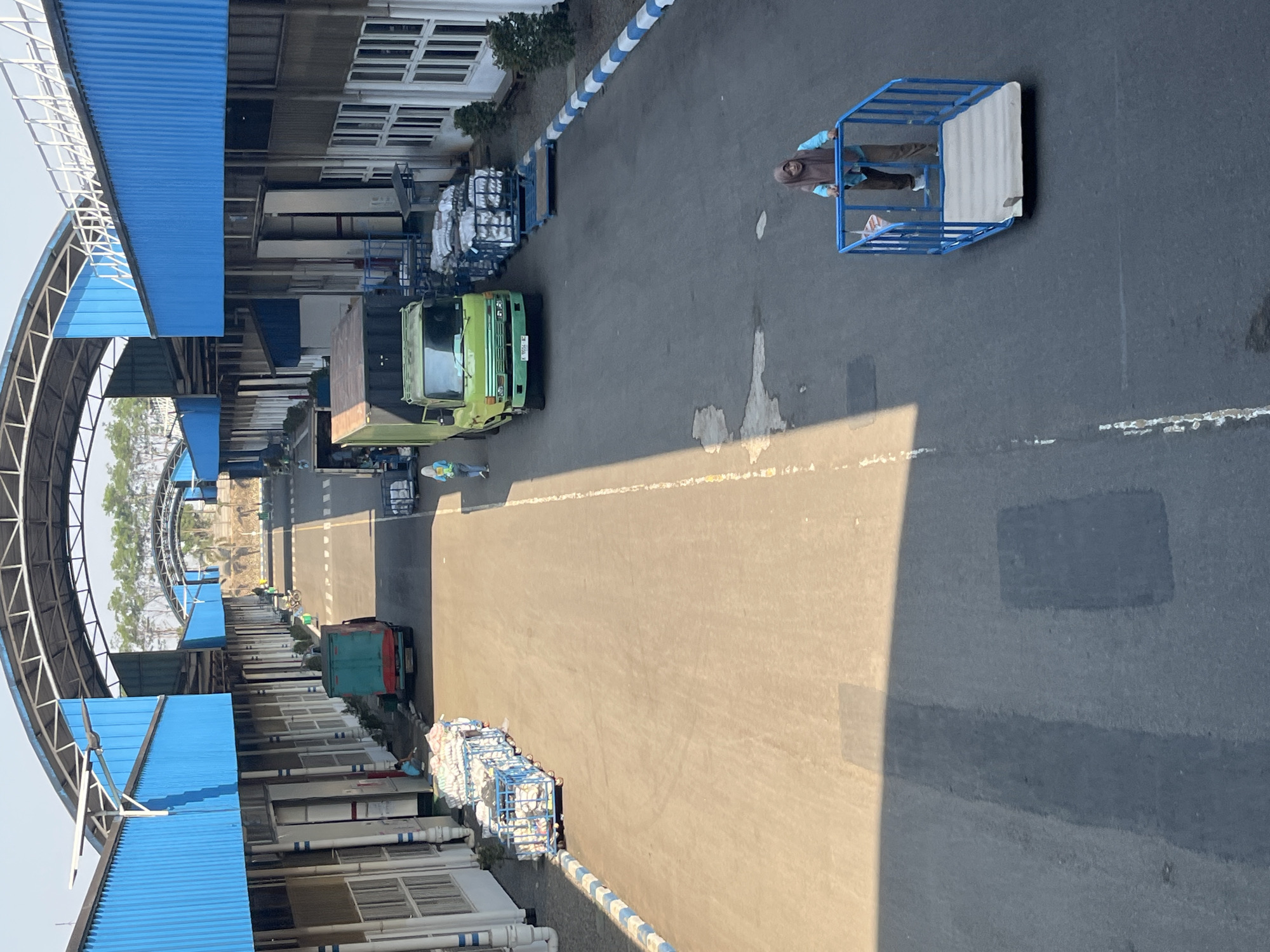 The image depicts an outdoor industrial area, possibly a loading or distribution center, in Pelang, Indonesia. The scene features a paved road flanked by large, blue-roofed buildings with numerous doors and windows. A woman wearing a hijab and a light blue top with brown pants is standing in a blue cage-like platform, likely a lift or cart, in the foreground on the right side of the image. Further down the road, a green truck is parked, and another truck is partially visible in the background. Several carts filled with white bagged items are lined up along the left side of the road. Another individual, possibly a worker, is walking near the green truck. The environment appears to be sunny, with shadows cast on the pavement. The overall impression is one of ongoing industrial activity.