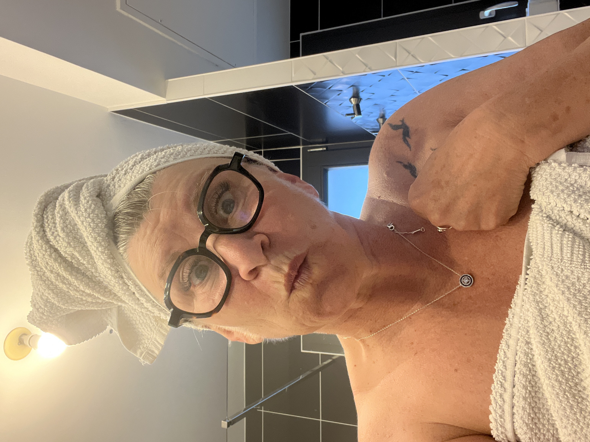 A woman with short gray hair is facing the camera in a bathroom. She is wearing black-rimmed glasses and a white towel wrapped around her head and body. She has a silver necklace with a round pendant. The bathroom has black and gray tiles, and there is a light fixture visible on the left. A tattoo is visible on her upper arm. The location of Chambœuf, France, cannot be confirmed from the image.