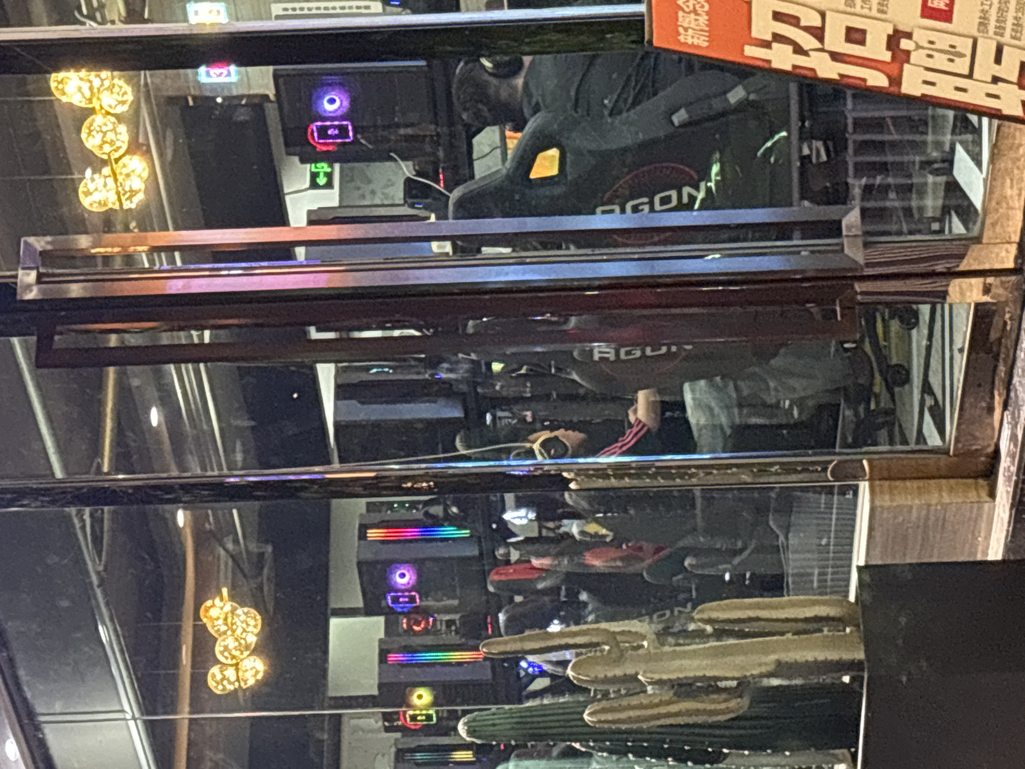 The image shows a reflection in a mirror of a gaming area, possibly an internet cafe, located in Nan'an District, China. In the reflection, several computer setups are visible with RGB lighting. A person is seen sitting in a gaming chair, possibly wearing headphones. There is a cactus near the mirror, and an advertisement or signage is partially visible at the top right corner.