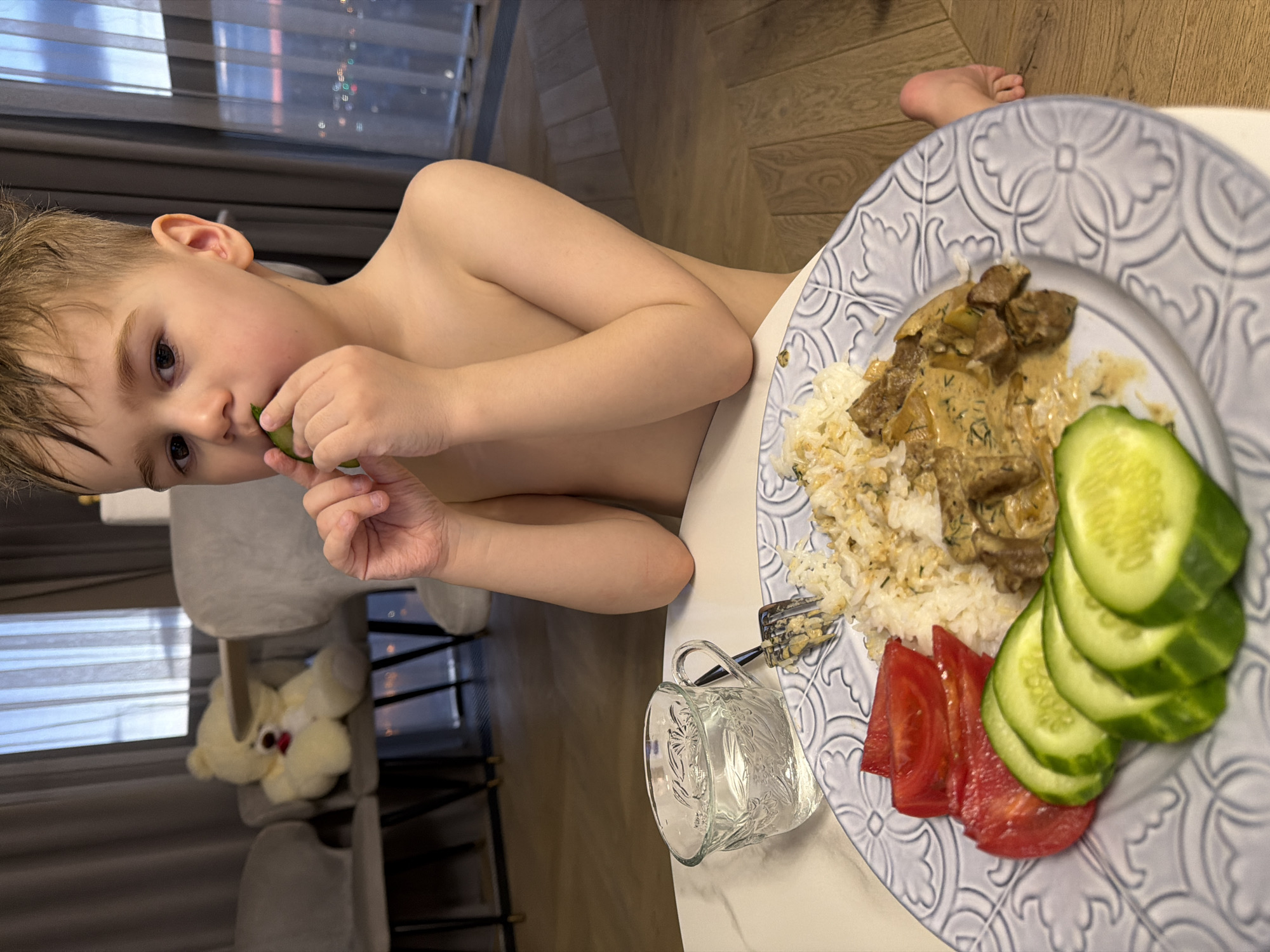 The image shows a young child, a boy with light brown hair, seated at a table during a meal. He is shirtless and is holding a slice of cucumber to his mouth, looking intently at it.
A large, round, decorative light blue-grey plate holds his meal, consisting of white rice next to chunks of meat covered in a creamy, herb-speckled sauce, possibly beef stroganoff. On the same plate, neatly arranged, are slices of fresh green cucumber and several red tomato wedges. A silver fork is visible on the plate, partially covered by the rice. Beside the plate, a clear glass cup with an embossed floral pattern rests on the table.
The setting is an indoor home environment, likely a dining area. In the background, a large window with horizontal blinds shows blurry lights, possibly city lights at night or reflections, suggesting the meal might be taking place in the evening. A grey upholstered chair is visible, and on it, a light-colored stuffed teddy bear with a red nose. The flooring is made of light-colored wood. The child appears focused on eating. No text is visible in the image. The scene depicts a common domestic activity, a child having dinner, consistent with a home in Krasnoyarsk, Russia.