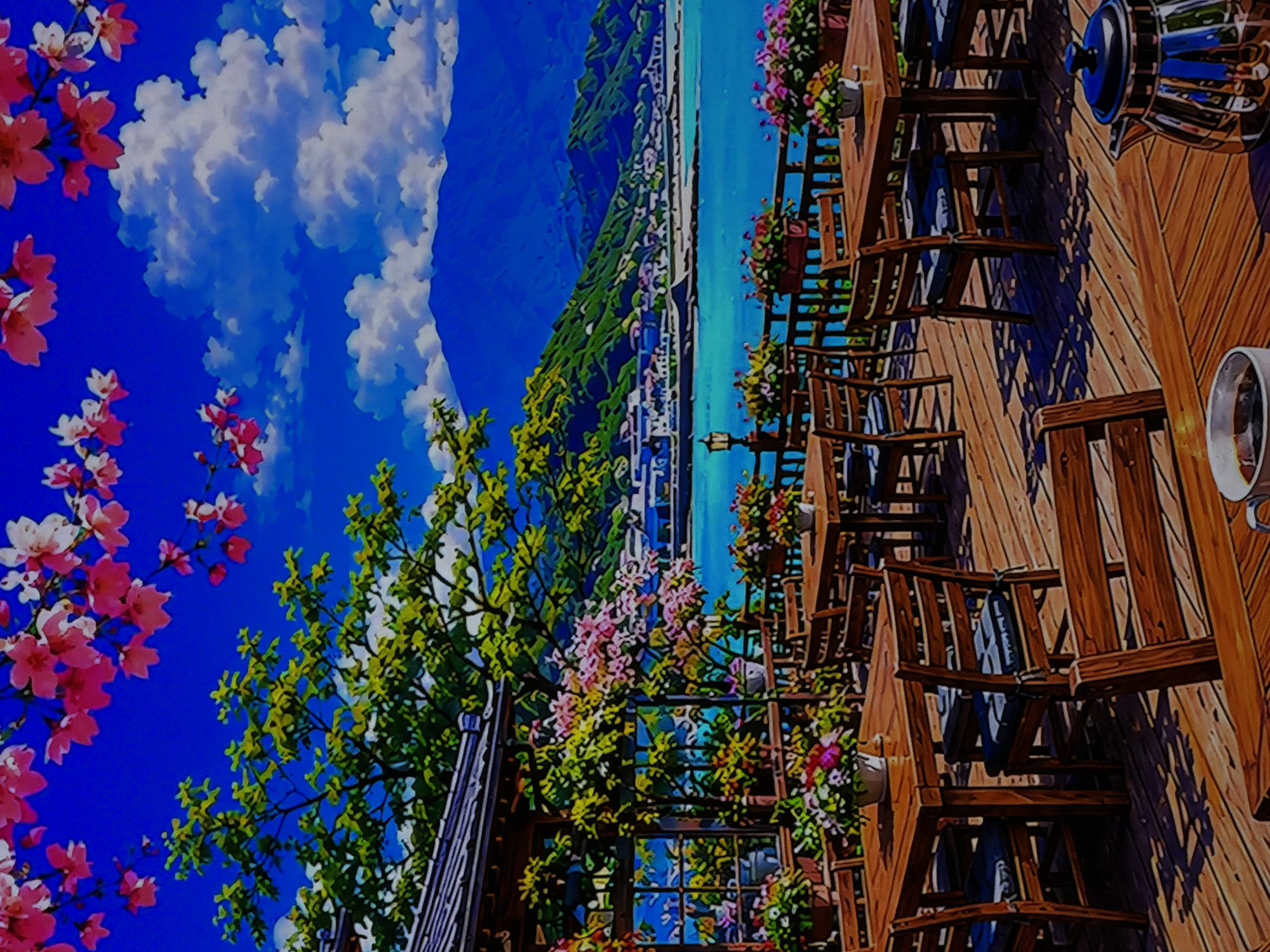 This is a vibrant, anime-style digital illustration depicting a picturesque outdoor cafe or restaurant with a scenic view. The image is rich in color, primarily dominated by blues, greens, and warm browns of the wood.
In the foreground, on the left side, branches laden with bright pink cherry blossoms frame the scene against a brilliant blue sky with fluffy white clouds. Below these, lush green foliage from other trees and what appears to be part of a dark-colored roof or eave are visible.
The midground features a rustic wooden deck, furnished with numerous empty wooden tables and chairs, arranged as if awaiting customers. Many tables and the deck railing are adorned with small potted plants and hanging baskets filled with colorful pink, purple, and white flowers, adding to the inviting atmosphere. Shadows cast on the deck and chairs indicate bright sunlight. On the far right, details such as a gleaming metallic teapot with a blue handle and lid, and a white ceramic mug, suggest that this is indeed a cafe setting.
Beyond the deck and flowers, a stunning panoramic view unfolds. A wide, tranquil blue body of water, possibly a sea or a large lake, stretches across the middle of the scene. Along the distant shore, a town or city with numerous buildings is nestled at the base of dramatic, green mountains that rise steeply into the background.
The scene is set during the daytime under clear, sunny skies, with scattered white cumulus clouds. No people are visible, contributing to a sense of peaceful serenity and inviting contemplation. The artistic style lends an idyllic and dreamlike quality to the entire setting. There is no visible text in the image.