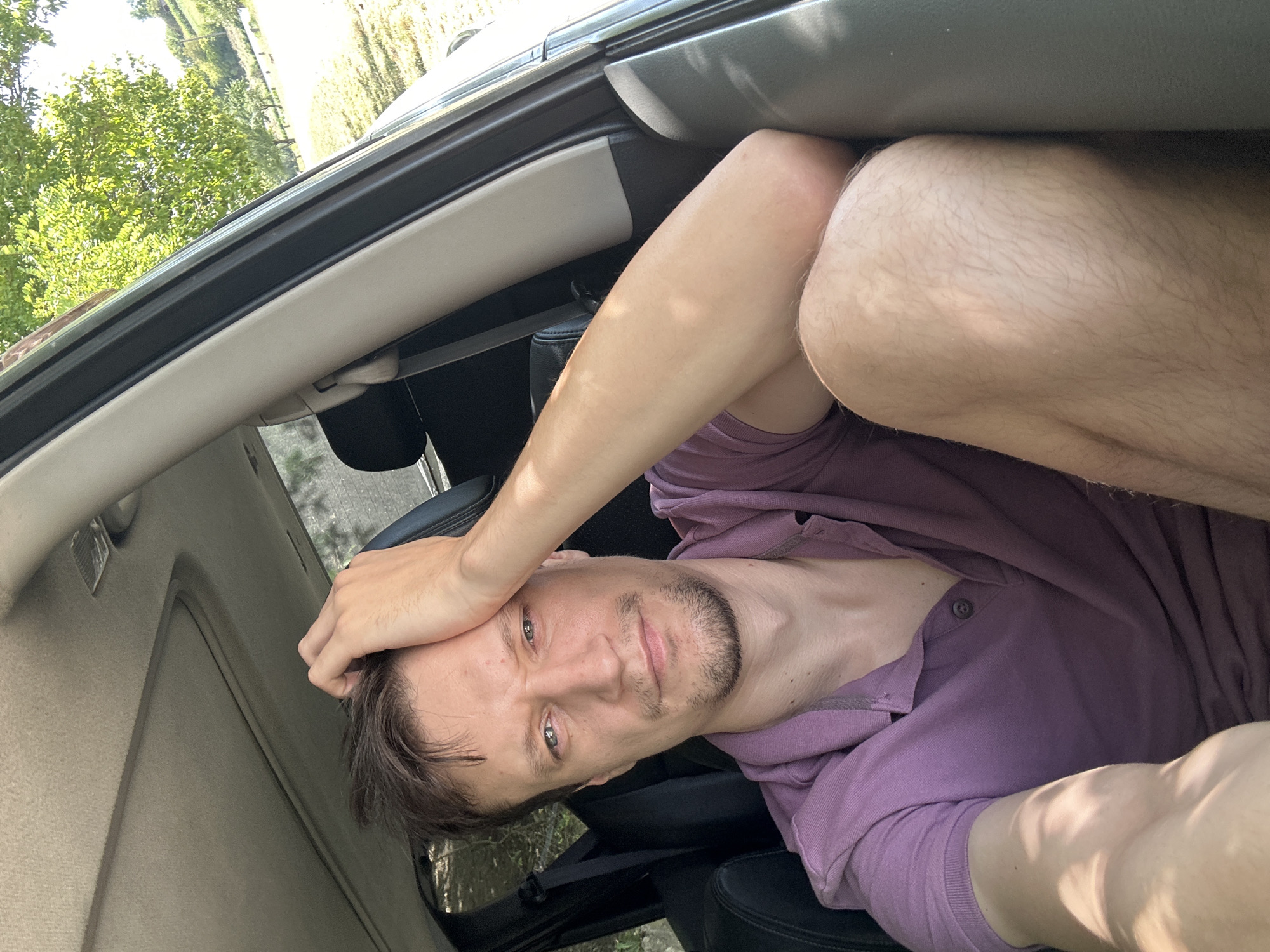 A medium close-up shot captures a man in his late 20s or early 30s sitting inside a vehicle. He has light brown hair, a light beard, and is wearing a purple polo shirt. The man's right hand is resting on his forehead, and his left arm is bent at the elbow, with his hand resting on his knee. The interior of the car is visible, including a sunroof and the car door. The background, seen through the open car door, shows a sunny, outdoor environment with trees and a hillside. The image appears to be a selfie taken by the man. The setting is outdoors, in or near Makiv, Ukraine.