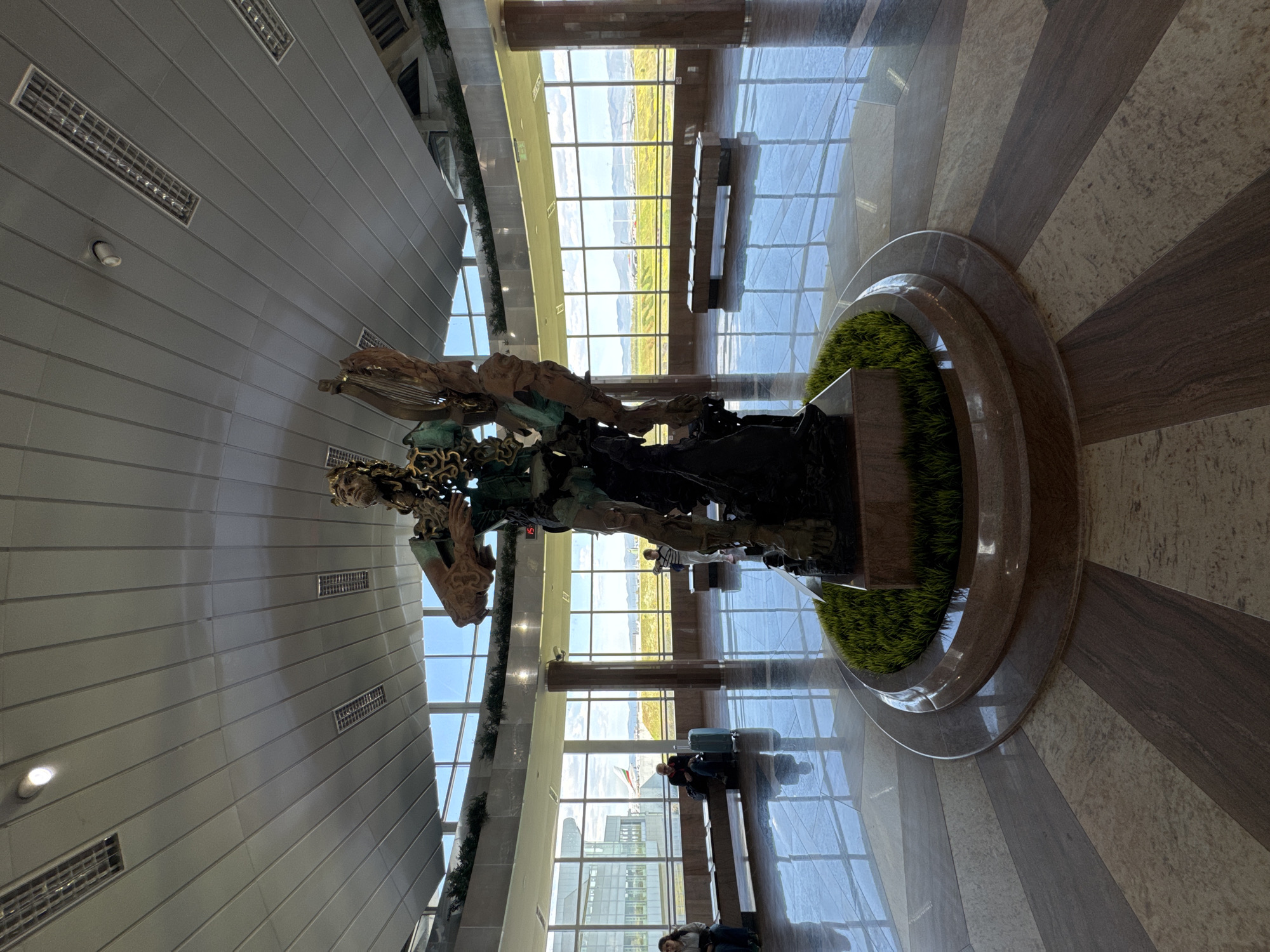 The image depicts a large, modern indoor space, likely a public building such as an airport terminal in Sofia, Bulgaria. The central focus is a complex, dark-colored statue or sculpture, possibly abstract or depicting multiple figures, resting on a circular base adorned with artificial green grass.
In the background, expansive, multi-paned windows dominate, offering a bright view of the outdoors. Through these windows, a vast, flat landscape with green and yellow fields is visible under a clear, sunny sky, with distant hills or mountains on the horizon. The bright daylight from outside illuminates the interior, creating reflections on the polished, striped floor, which features alternating bands of brown and beige/cream tones.
Several individuals are present in the scene. A person is clearly visible sitting on a bench in the bottom right, next to a turquoise and a dark suitcase, suggesting they are waiting. Other figures are scattered further back near the windows, some seated, some standing. The overall activity appears to be people waiting or passing through.
The architecture is contemporary, with a high ceiling featuring light grey panels and rectangular ventilation or lighting fixtures. A small, circular ceiling light is also visible. Interior structures include pillars and possibly artificial greenery lining some edges. A small red digital display on a pillar near the statue shows the number "15". The atmosphere is bright, clean, and orderly.