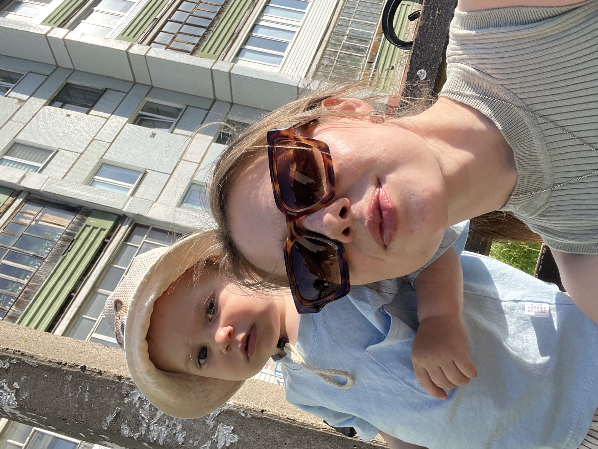 The image shows a woman and a young child taking a selfie. The woman is wearing large tortoiseshell sunglasses and a light-colored ribbed top. The child is wearing a light blue shirt and a beige sun hat. The background features a multi-story building with a gray facade and many windows. Based on the building style, the location can be presumed to be Chaikovsky, Russia. They are outside near a concrete ledge. The child looks at the camera, while the woman's face is partially obscured by the sunglasses.