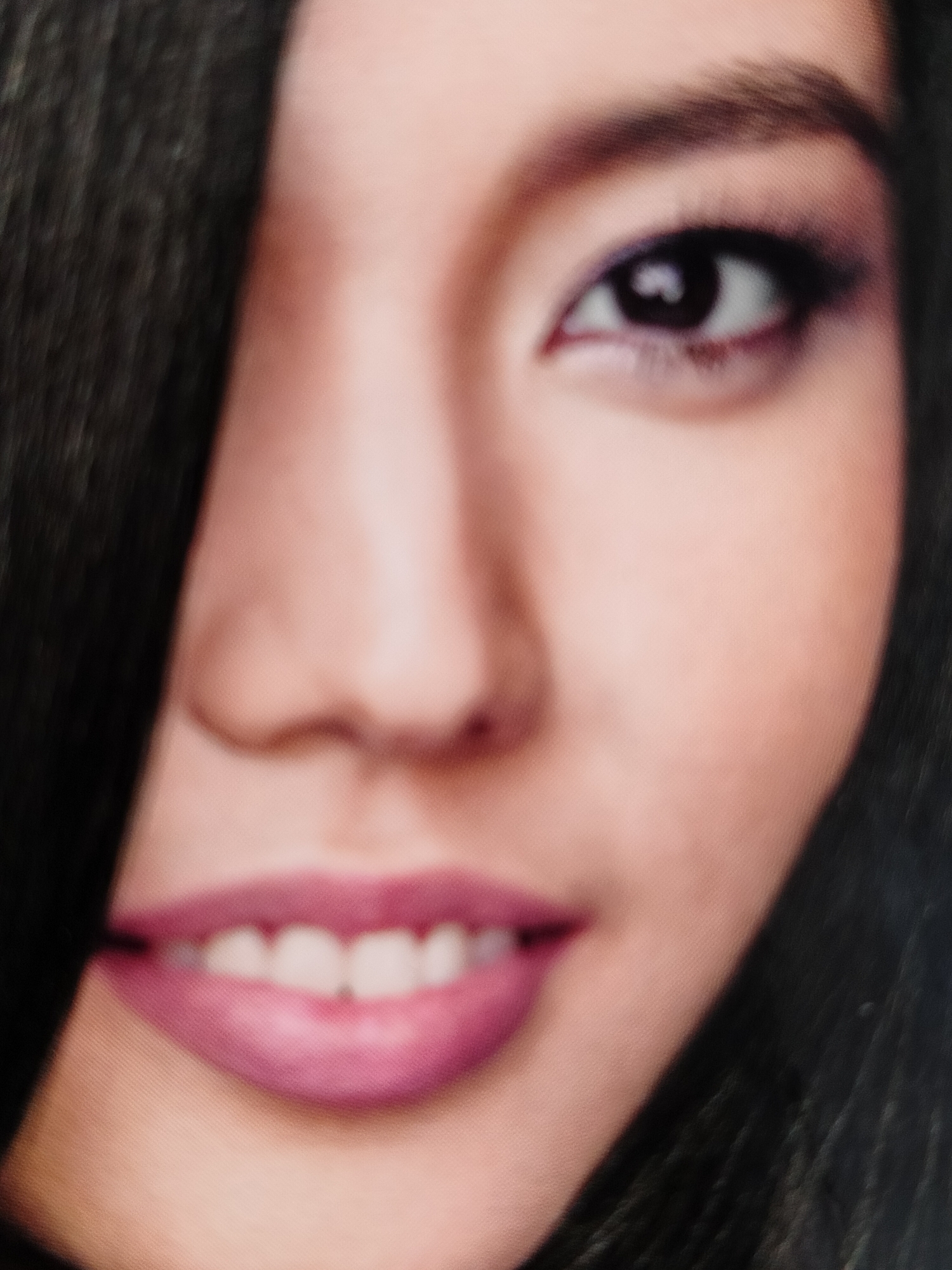 The media file is a close-up, portrait-style image of a person's face, focusing on their eye, nose, and mouth area. The subject appears to be a young woman with dark hair, which is partially obscuring her left side. Her right eye is visible, adorned with dark eyeshadow and eyeliner, and her eyebrow is well-defined. She has a light skin tone. Her mouth is open, revealing a set of white teeth and lips colored with a vibrant pink lipstick. The lighting suggests it's likely indoors or in a controlled environment, as there are no discernible shadows indicative of natural daylight. The overall mood conveyed is one of beauty and possibly confidence, due to the makeup and open smile. The image quality is somewhat low, with a noticeable graininess. There is no visible text in the image. The location context of Novosibirsk, Russia, does not provide any specific visual cues within the image itself.