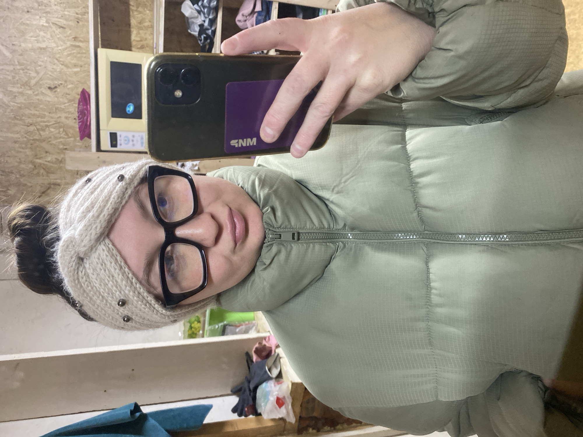 A woman is taking a selfie indoors. She wears a light green puffer jacket, a knitted headband, and black-rimmed glasses. She is holding a smartphone with a purple card attached to the back, obscuring part of the phone. The background shows a plywood wall and cluttered shelves. The location might be Chaikovsky, Russia, but this cannot be confirmed from the image alone.