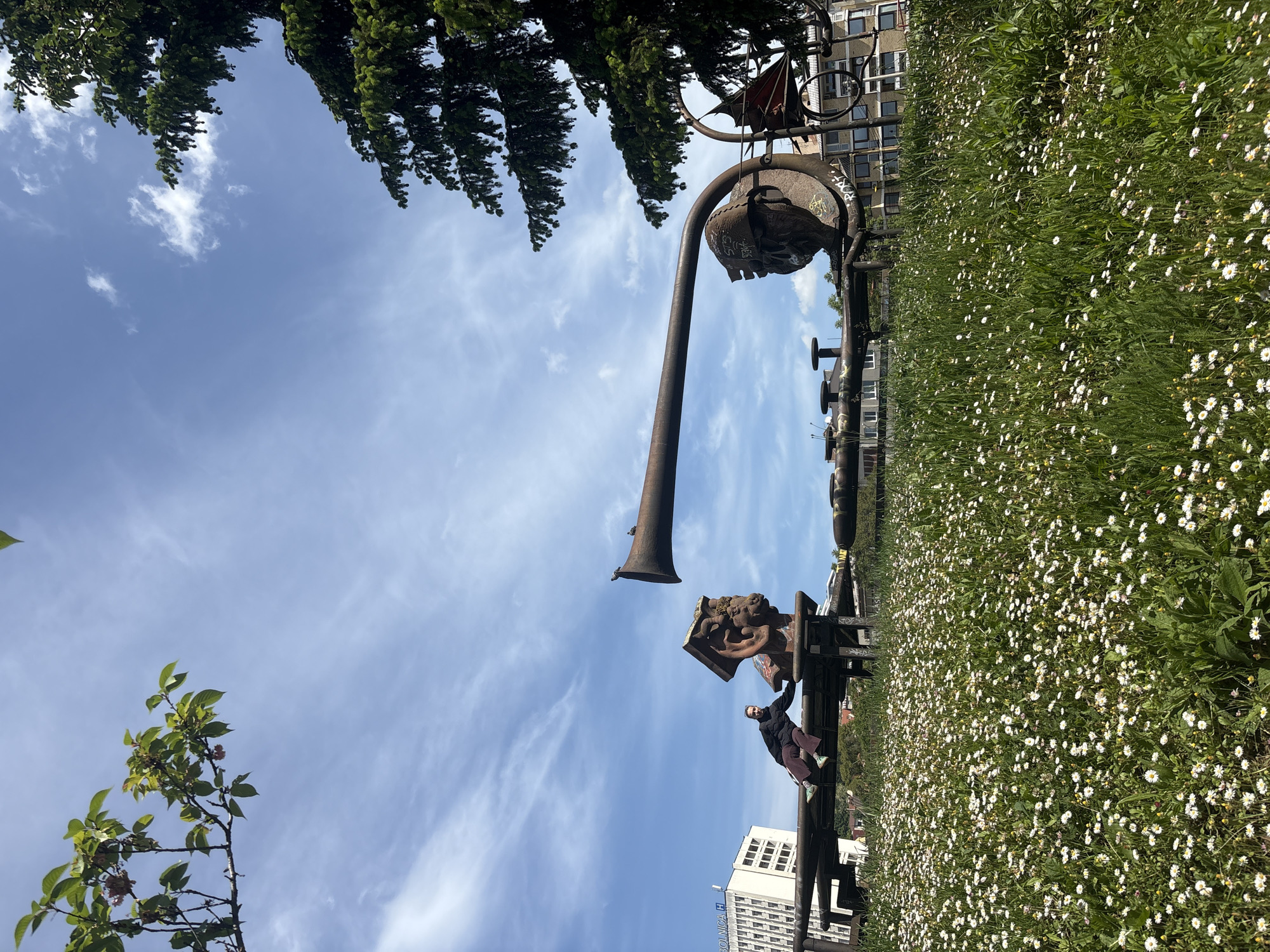 The image shows a metal sculpture in Sarajevo, Bosnia and Herzegovina. The sculpture features a large, rusted metal face with an elongated nose resembling a trumpet. A person is seated on a bench that is part of the sculpture. The background includes a field of daisies, trees, buildings, and a blue sky with scattered clouds. The sculpture is located in an outdoor park or green space within the city.