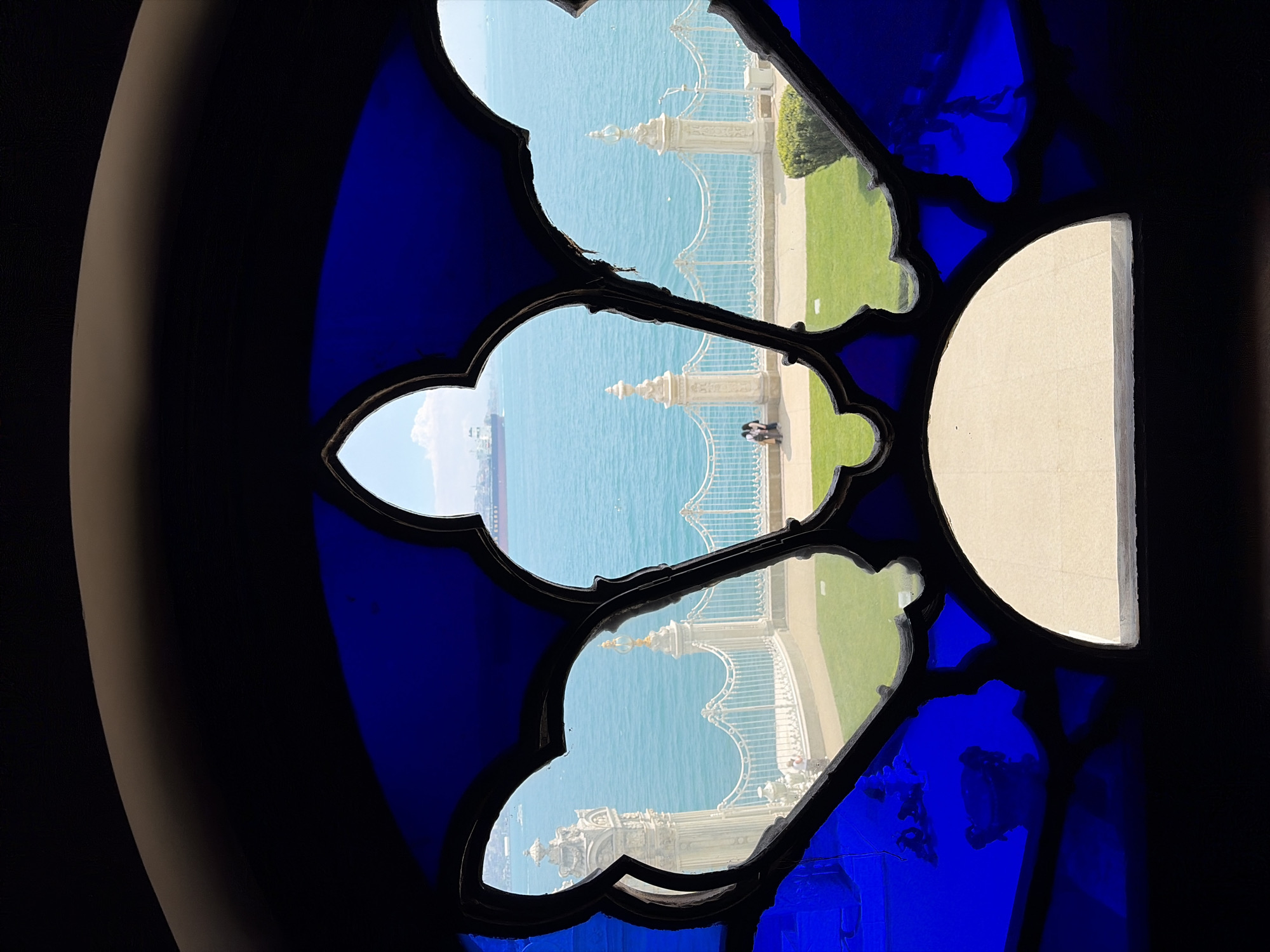 The image shows a view of the Bosphorus Strait in Beşiktaş, Turkey, framed by a blue stained-glass window. The window has a distinctive quatrefoil design, and a section of it is missing on the right side, replaced by a plain glass pane. Through the openings, a portion of a waterfront pathway with a white wrought-iron railing is visible. The pathway is bordered by grass and the sea. Two individuals are standing on the pathway in the central opening. A ship is visible in the distance of the central opening.