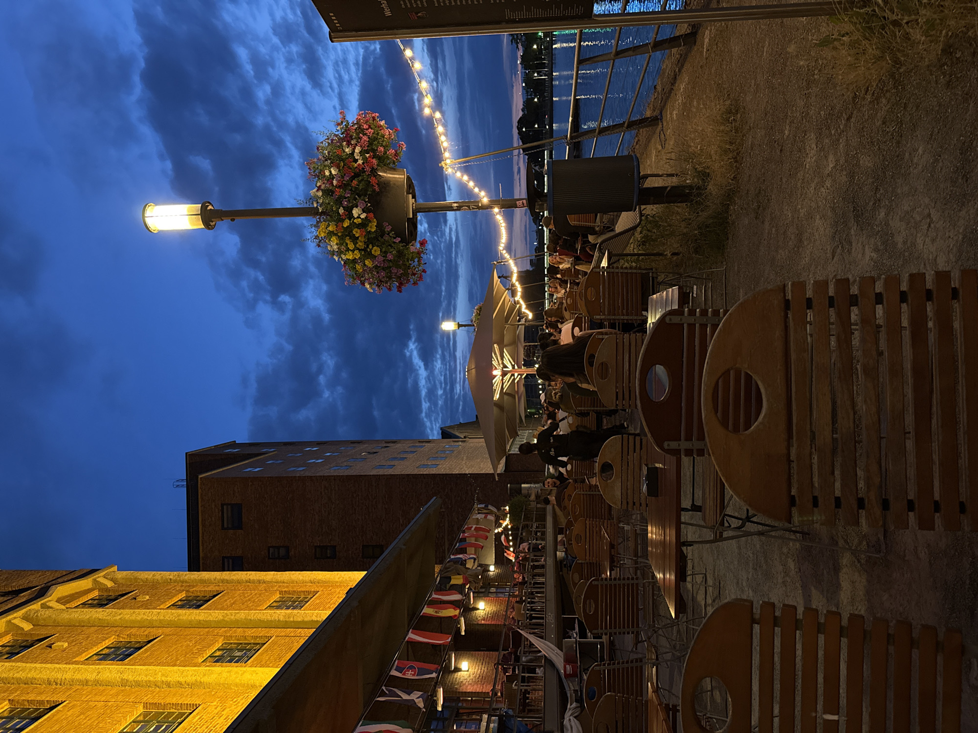 This image captures an outdoor seating area of a restaurant or cafe at dusk in the city of Duisburg, Germany. The scene is situated along a waterfront, with a body of water visible on the right side of the frame.
In the foreground, rows of empty wooden tables and chairs are arranged, suggesting a dining or lounge area. The tables are made of wood with metal legs, and the chairs are also wooden with slatted backs and seats. Some ashtrays are visible on the tables.
Behind the empty seating area, a covered patio area is visible, lined with tables and chairs where patrons are seated. People are dining and socializing in this area, some under umbrellas or awnings. A waiter or staff member wearing a dark uniform is visible walking among the tables.
The background features a large, yellow brick building, likely an industrial or commercial structure, illuminated by warm yellow lights. To the right of this building is another taller brick building. A lamppost with a flower basket at its base and a lit lamp stands prominently in the middle ground, adorned with a string of white lights that stretch across the scene.
The sky is a deep blue, indicative of twilight, with dark clouds creating a dramatic backdrop. The waterfront is calm, reflecting the ambient light. A railing separates the seating area from the water. A dark trash receptacle is positioned near the lamppost.
A sign is mounted on a pole to the far right, with text that is partially legible and appears to be a menu or information board. Flags of various countries are hung along the covered patio area. The overall impression is a lively evening scene at a waterfront establishment.