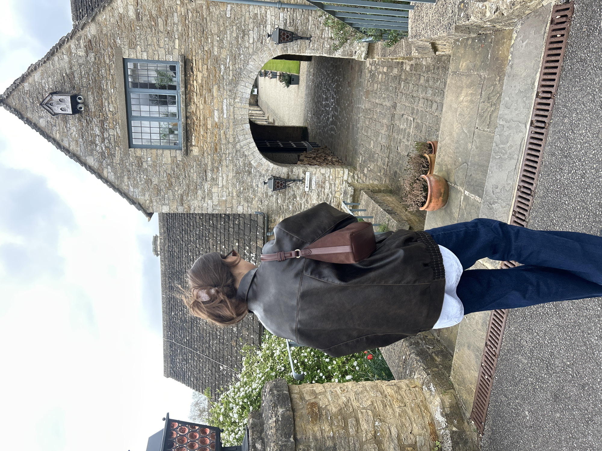 A young woman wearing a dark brown jacket and blue jeans stands on the side of a street in Malmesbury, United Kingdom. She has a brown purse on her shoulder and is looking at the stone building and archway in front of her. The building is made of light-colored stone and has a window and a decorative crest above it. A sign reading "Reception" is on the archway. The street is paved and has a metal drainage grate. Greenery and flowers grow along the walls.