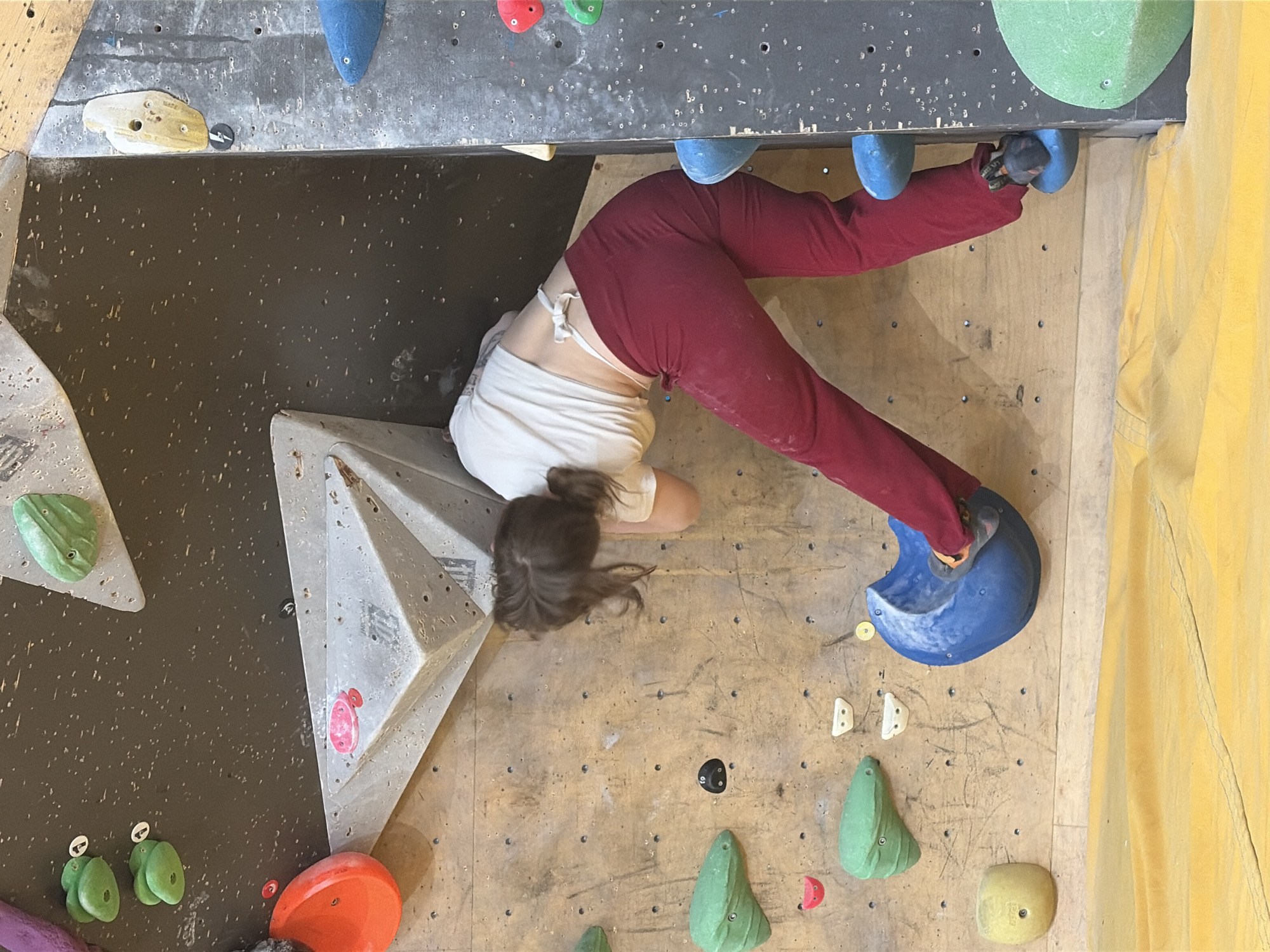 A person is climbing a bouldering wall. The person is wearing a white t-shirt and red pants. The person is positioned in the middle of the frame, facing away from the camera. Their body is angled to the left, with their right leg extended. Their left foot is placed on a blue climbing hold, and their right foot is on another blue climbing hold. The climbing wall is made of wood and has various colorful climbing holds attached to it. The background is a dark wall with speckles. There is a yellow mat on the floor in front of the climbing wall. There is no information in the image that suggests it is located in Moscow, Russia.
