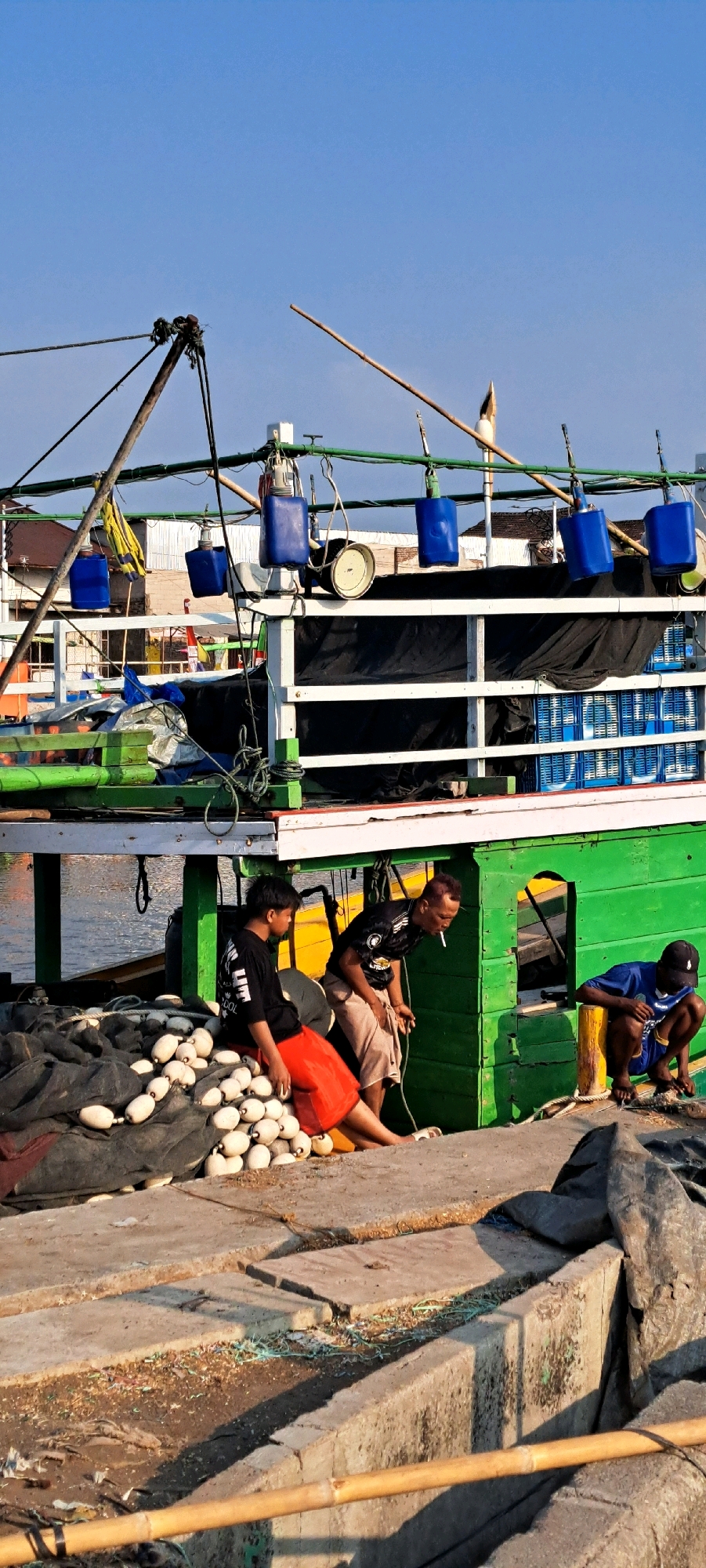 This image depicts a scene at a harbor in Pasuruan, Indonesia, with a focus on a fishing boat and its crew. The boat, painted green and white, is docked alongside a concrete pier. A pile of fishing nets with white buoys is situated on the pier, next to two men.
One man, wearing a black t-shirt and red shorts, is seated on the fishing nets. Another man, in a black t-shirt with a white logo and light brown shorts, is standing and appears to be talking or gesturing. A third man, wearing a blue t-shirt and dark shorts, is crouched down further along the pier.
Above the boat, the sky is clear and blue. Various equipment, including blue plastic containers and a winch, are visible attached to the boat's rigging. In the background, buildings and other boats can be seen. The overall impression is one of activity or preparation related to fishing operations.