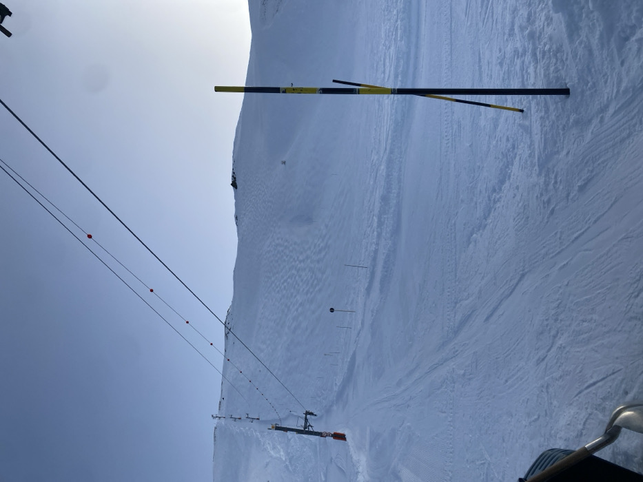 The image displays a snow-covered ski slope in La Salle-les-Alpes, France, under a clear, pale blue sky. The terrain is primarily white snow, exhibiting varied textures.
On the left side of the mid-ground, a section of the ski slope shows distinct parallel corduroy grooming patterns, indicative of recent snowcat activity. To the right, the snow surface appears less uniformly groomed, presenting undulating patterns and scattered tracks.
A ski lift system extends diagonally from the mid-ground up the left side of the slope. This system comprises multiple dark-colored pylons supporting overhead cables. Attached to these cables, at regular intervals, are small, red spherical marker balls.
In the lower right foreground, a thick, vertically oriented black pole with alternating yellow and black stripes is embedded in the snow. A second, thinner pole with a similar yellow and black stripe pattern leans against or is positioned immediately behind the primary pole. Further up the slope, several slender dark poles, some with indistinct markers, are sparsely distributed. A circular dark sign mounted on a pole is visible in the mid-ground, slightly left of center.
The background features steep, snow-clad mountain slopes, with dark rocky outcrops visible along the higher ridges. The scene is illuminated by bright daylight, with a minor atmospheric haze or glare present in the upper right portion of the sky. No individuals are present in the frame. A metallic object, consistent with a shovel blade and part of a wooden handle, is partially visible in the extreme lower-left corner.