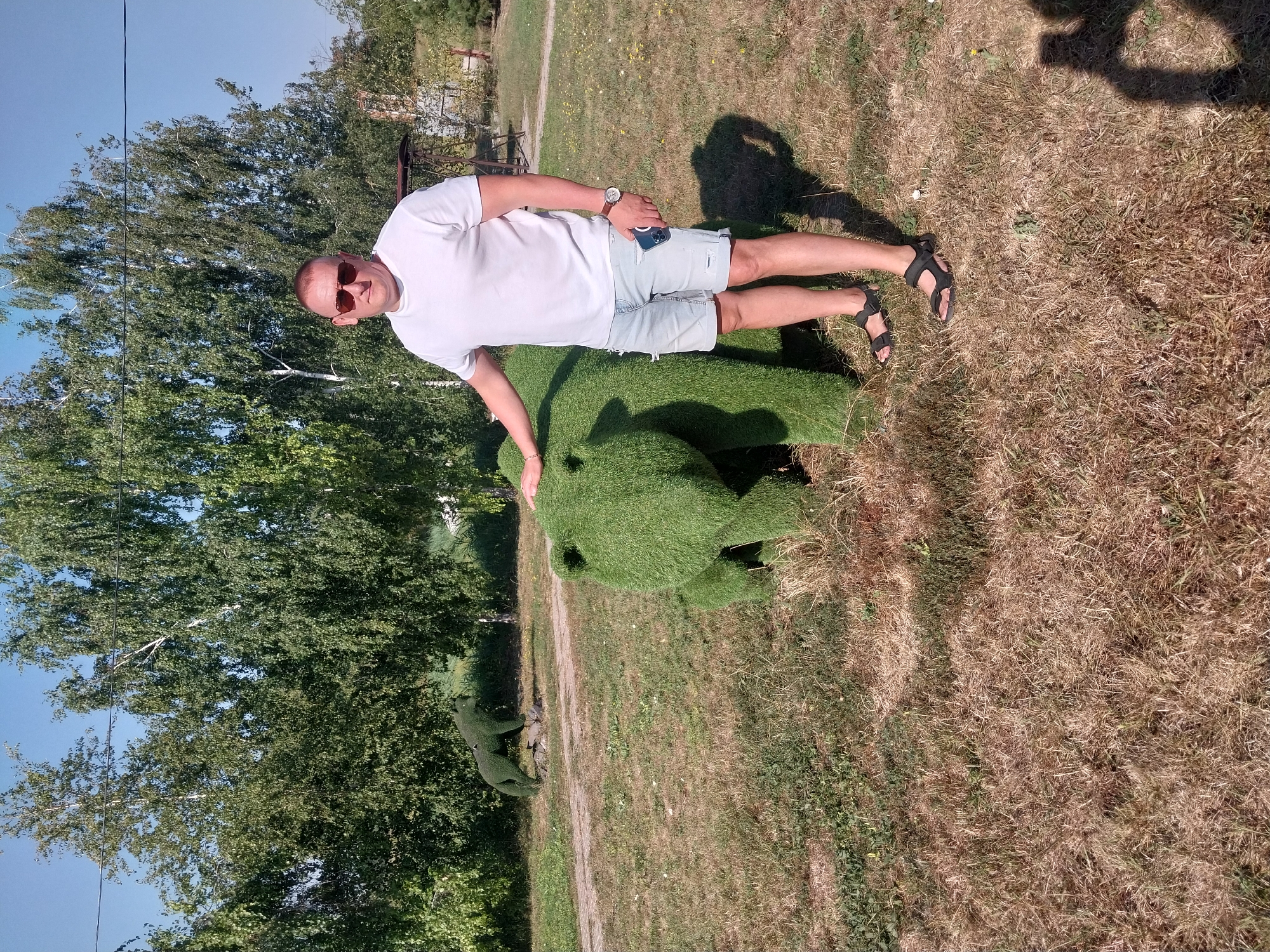 In a park setting in Petrykivka, Ukraine, a man stands next to a topiary sculpture of a bear. The man is wearing a white t-shirt, light-colored ripped denim shorts, and black sandals. He has on sunglasses and a watch. In the background, there are trees and a second topiary sculpture of a wolf. The ground is covered with dry grass.