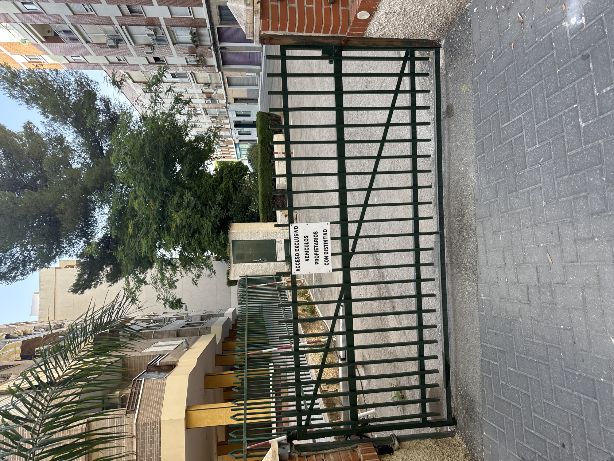 The image depicts a closed green metal gate in front of what appears to be a residential complex. A sign attached to the gate reads "ACCESO EXCLUSIVO VEHICULOS PROPIETARIOS CON DISTINTIVO," indicating exclusive access for resident vehicles with a permit. The gate is situated on a paved walkway made of interlocking gray bricks. Behind the gate, a dirt area is visible, with some sparse vegetation. In the background, there are multi-story apartment buildings, one with a yellow facade and another made of red brick. A tall tree with dense green foliage stands prominently behind the gate. The scene appears to be in an urban setting, likely a residential area. The lighting suggests it is daytime, with natural light illuminating the scene. There are no people visible in the image.