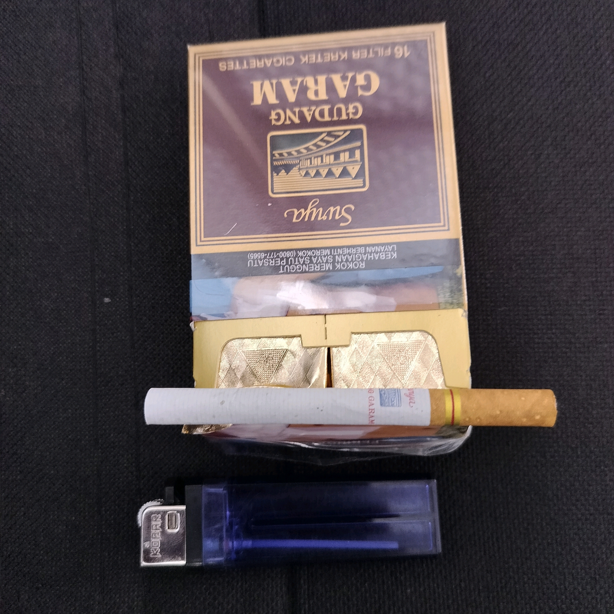 The image shows a dark, textured surface, likely a tablecloth or fabric. In the center of the frame, there is an open pack of Gudang Garam Surya cigarettes, with the cigarettes partially visible. One cigarette is lying horizontally across the top of the open pack, and its filter end is on the right side of the image. Below the cigarette pack, a blue disposable lighter is placed horizontally. The lighter has a metallic striking mechanism with the word "KORR" embossed on it. The scene appears to be a still life arrangement of smoking paraphernalia on a dark background. The image does not provide any information about the location in the city of Sleman, Indonesia.