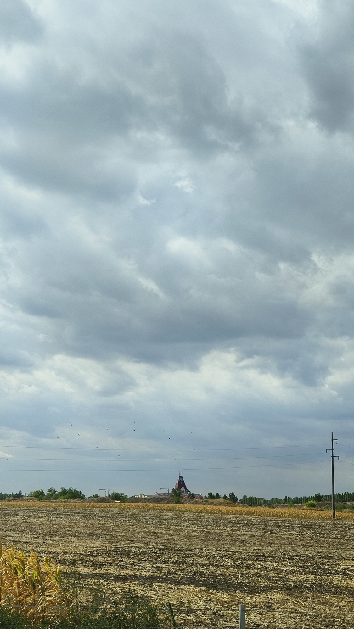 The image shows a wide shot of a rural landscape under a dramatic, cloudy sky. In the foreground, a field of harvested crops, likely corn stalks, is visible, with some green vegetation in the lower left corner. In the middle ground, a row of trees separates the harvested field from a slightly elevated area where a distinctive, cone-shaped building with a red roof stands. This building appears to be a unique architectural structure, possibly a landmark or tourist attraction. A power line stretches across the frame horizontally in front of the building and trees, supported by a utility pole on the right side of the image. The sky is filled with heavy, gray clouds, suggesting an overcast or potentially stormy day. The scene is set in or near Palić, Serbia, as indicated by the presence of this notable structure. There are no people visible in the image, and the overall atmosphere is serene yet imposing due to the dramatic sky.