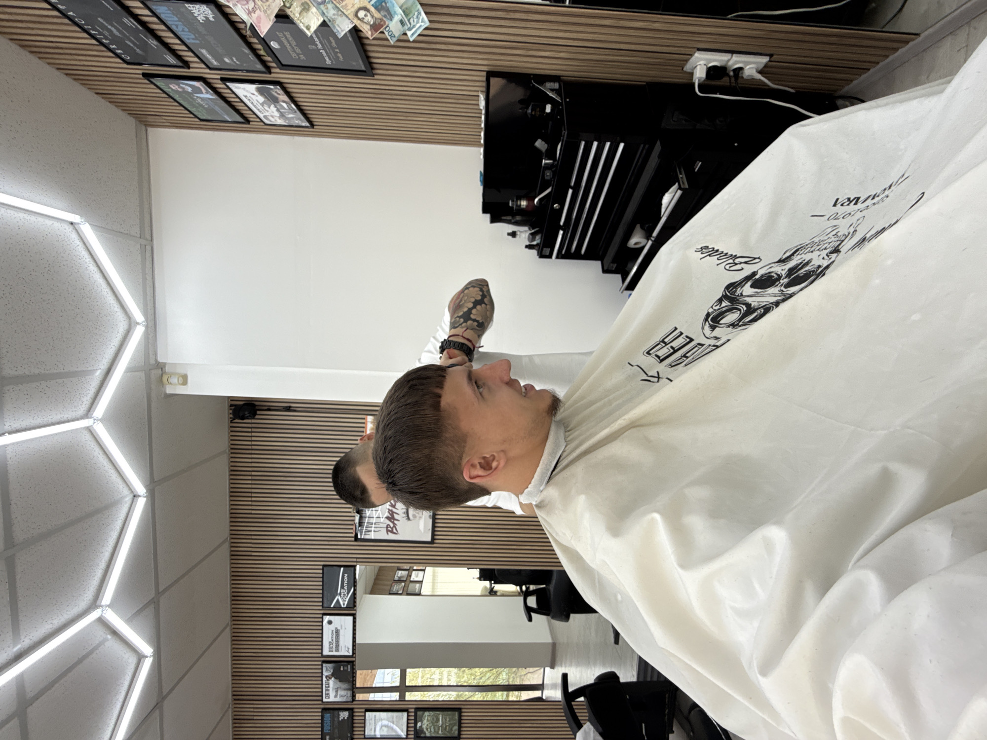 Here is a detailed description of the image:
**Scene:** The image shows the interior of a barbershop.
**People:** Two men are visible. One is seated and covered with a white barber's cape, indicating he is a customer receiving a haircut. The other man is partially visible and appears to be the barber, as he's standing and has his arm raised.
**Objects:** Key objects include a black barber's tool chest, various certificates and framed pictures on the wall, a mirror, barber's chairs, and cash displayed on the wall. A white barber's cape is draped over the seated customer.
**Actions:** The barber seems to be in the process of cutting the customer's hair.
**Interactions:** The image captures the interaction between the barber and the customer during a haircut appointment.
**Environment:** The barbershop is well-lit, with a modern and clean appearance. The walls are decorated with certificates, pictures, and cash. The floor seems to be tile or another smooth material. There is an emphasis on professionalism and quality service.
**Location:** The certificates in the image indicate the city of Sofia, Bulgaria.