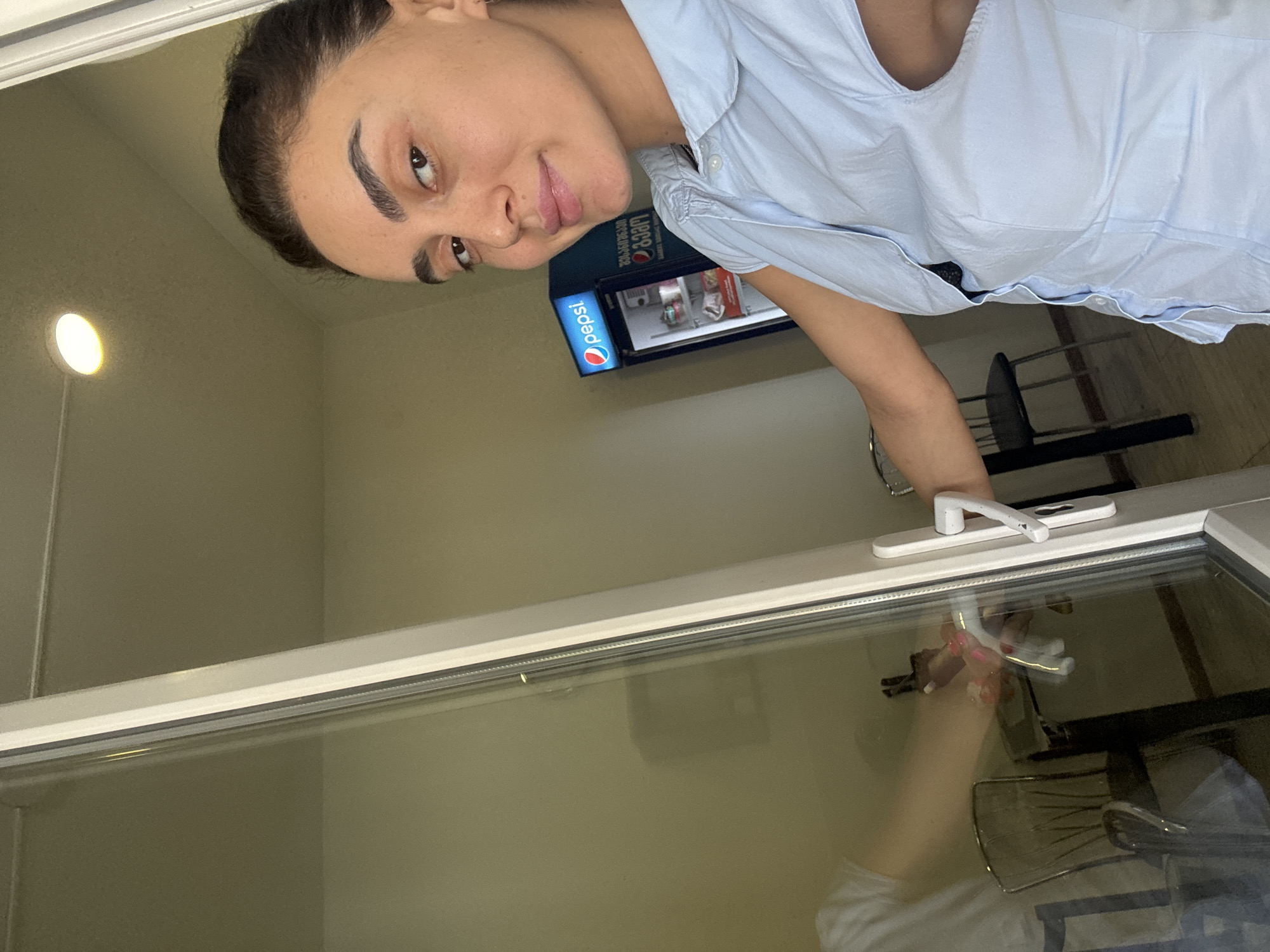 A woman with dark hair and prominent eyebrows is taking a selfie in a room that appears to be a hotel room. She is wearing a light blue short-sleeved collared shirt and a black bra. To her right, a small Pepsi refrigerator is visible. The room has light-colored walls and a ceiling with a circular light fixture. A glass door with a white handle is in the foreground, reflecting some of the room's interior. The woman's left arm is extended and holding the door handle. The reflection in the glass shows a table with chairs and what appears to be a pink toy. The scene suggests the woman is about to exit the room or has just entered it. Based on visual cues like the Pepsi refrigerator and the interior decor, this setting is consistent with a hotel room in Tbilisi, Georgia.