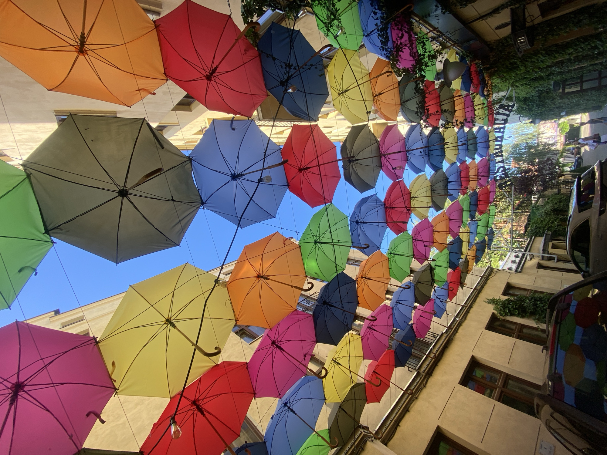 The image depicts a street scene in Łódź, Poland, featuring an overhead canopy of numerous colorful umbrellas. The umbrellas are suspended across the street, creating a vibrant and festive atmosphere. They are arranged in multiple layers, varying in color, including pink, yellow, green, orange, blue, red, and grey. The buildings flanking the street are visible, characterized by their light-colored facades and windows. In the foreground, two cars are parked. The right side of the street is adorned with lush green ivy growing on a building. In the distance, an archway with the word "RESTAURACJA" is visible, indicating a restaurant. Several people are present in the image, walking on the street. The sky is clear and blue, indicating a sunny day.