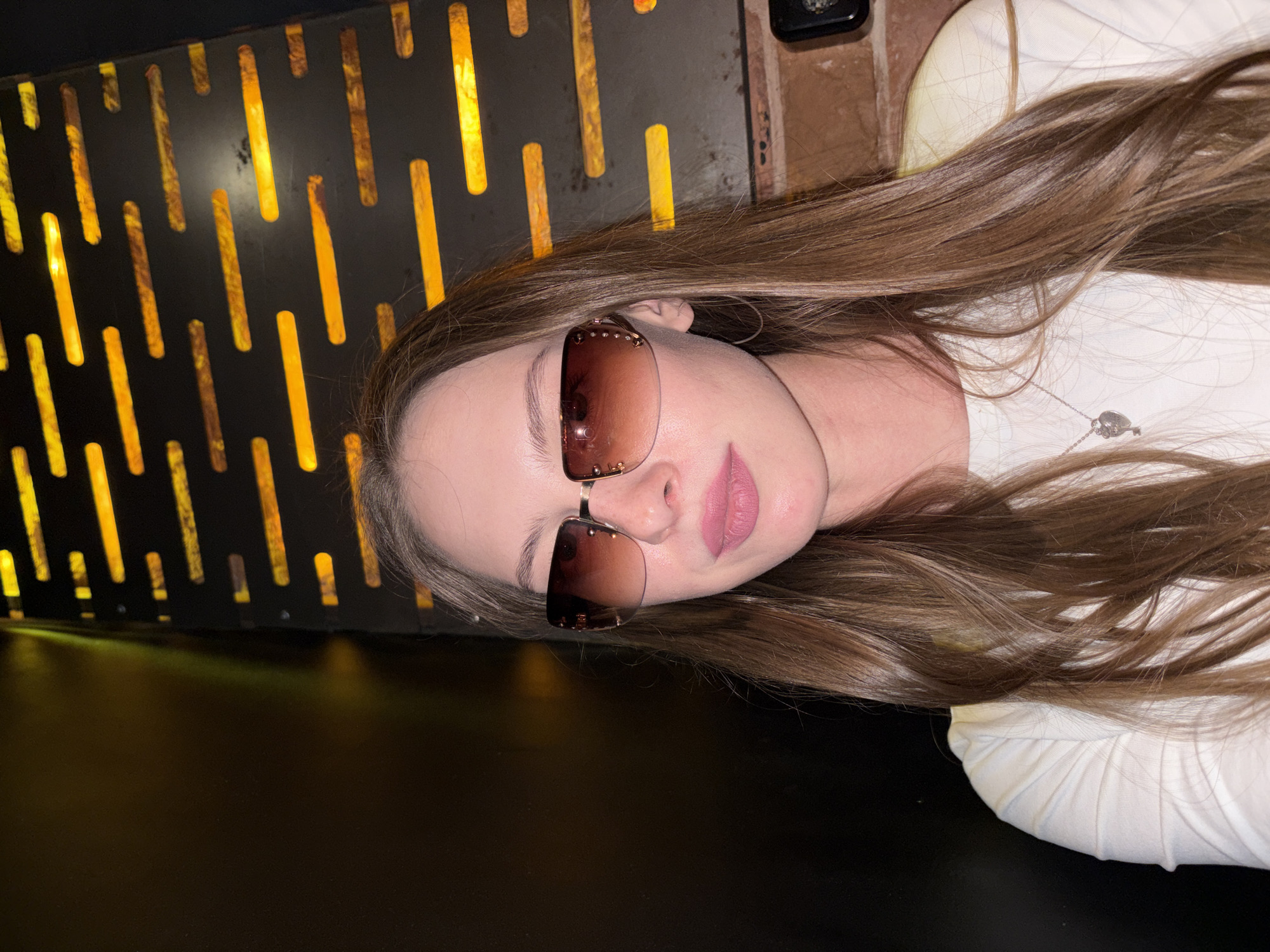A young woman with long, light brown hair is pictured wearing sunglasses and a white shirt. She is indoors, with a dark, textured wall with yellow lights behind her. A brick wall is visible to the right of the image. The overall lighting suggests an evening or dimly lit indoor setting. This image does not provide any information to determine its location within the city of Perm, Russia.