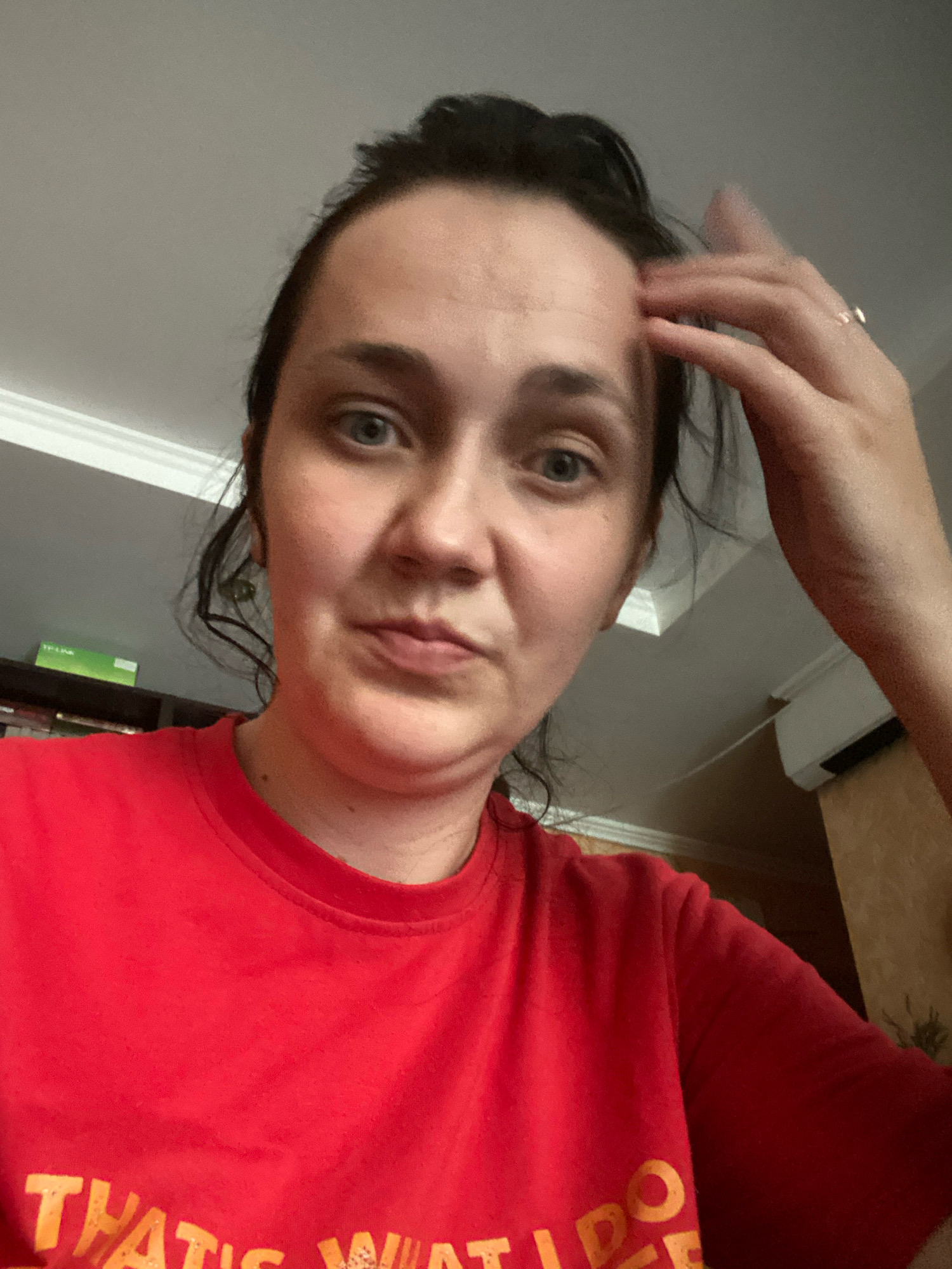 A medium shot captures a woman with dark hair pulled back in a bun and loose strands framing her face. She is wearing a bright red t-shirt with yellow lettering that is partially visible. Her right hand is raised, touching her forehead and temple, and a ring is visible on her ring finger. The woman's gaze is directed slightly off-camera. The background appears to be indoors, with a white ceiling and crown molding visible at the top. To the left, a shelf with some books and a green box is partially visible. On the right, an air conditioning unit is mounted on the wall. The overall lighting suggests an indoor environment.