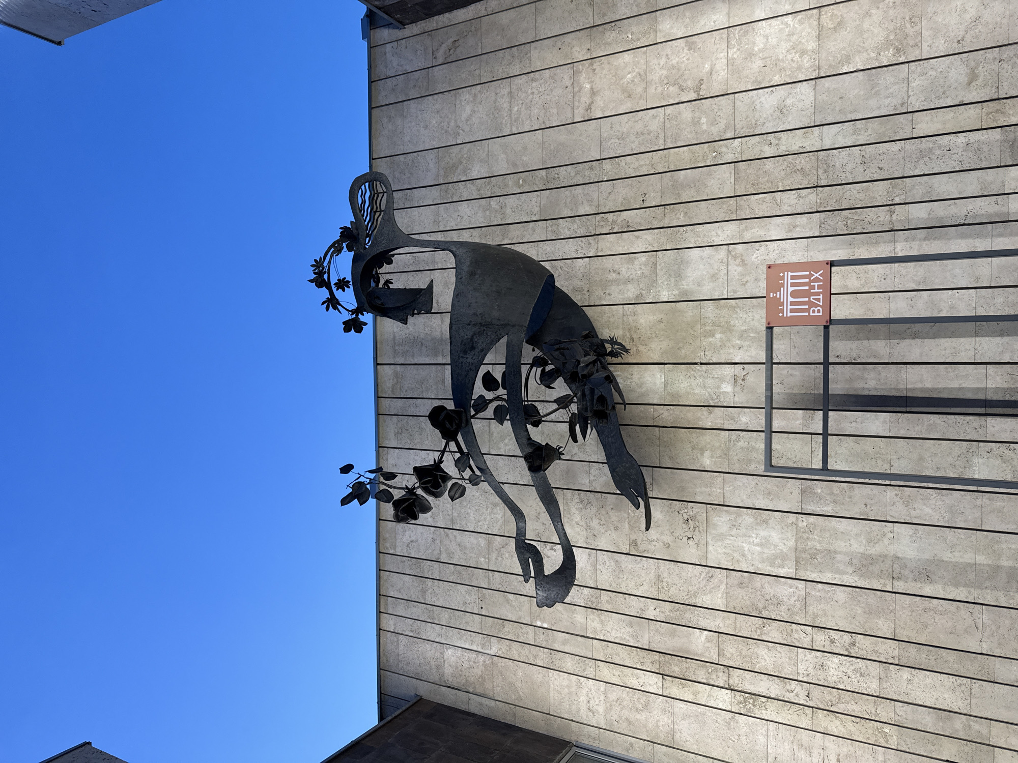 The image shows a close-up, low-angle view of a large, dark metal sculpture mounted on a beige tiled building wall under a clear blue sky. The sculpture depicts a stylized human figure with flowing lines, resembling a bird or a dancer, adorned with metal roses and leaves. The figure's head is in profile, with a wreath of flowers around it, and the body extends in a dynamic pose. Below the sculpture, there is a dark metal railing and a small, square sign with white text and graphics on a reddish-brown background. The text on the sign reads "ВДНХ" in Cyrillic letters, and features a graphic of arches. This indicates the location is at VDNKh (Exhibition of Achievements of National Economy) in Moscow, Russia. The building facade is made of vertical, rectangular beige tiles. The lighting suggests it is daytime, with shadows cast by the sculpture and railing.