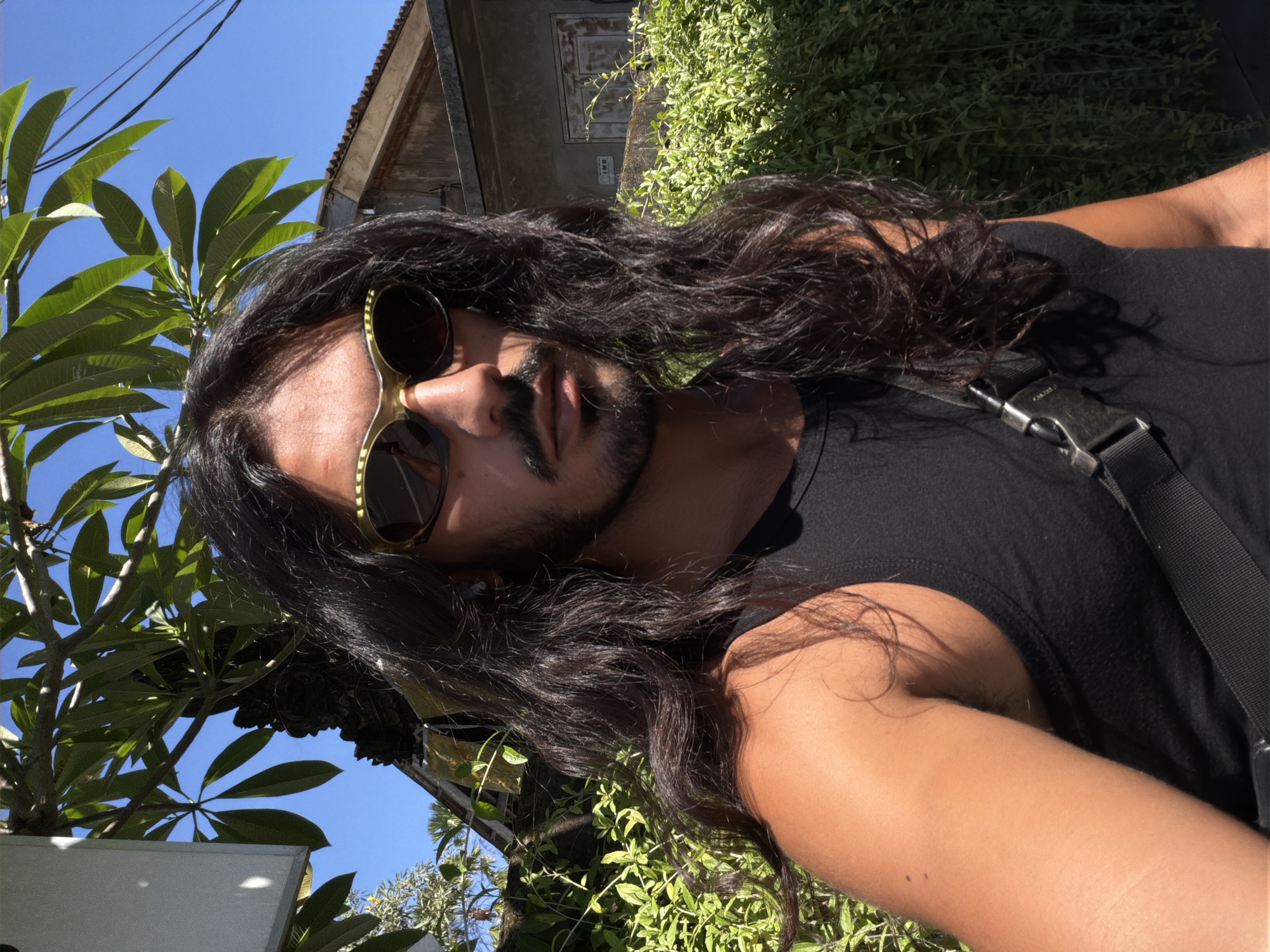 The image depicts a man with long, dark hair and a mustache, wearing gold-framed sunglasses and a black tank top. He is positioned outdoors, with a building and green foliage visible in the background. The blue sky suggests a sunny day. Based on the location provided (North Kuta, Indonesia), the architectural style of the building and the tropical plants are consistent with that region. A strap with a buckle is visible across his chest, possibly for a bag. The image appears to be taken from a low angle, tilting upwards towards the man.