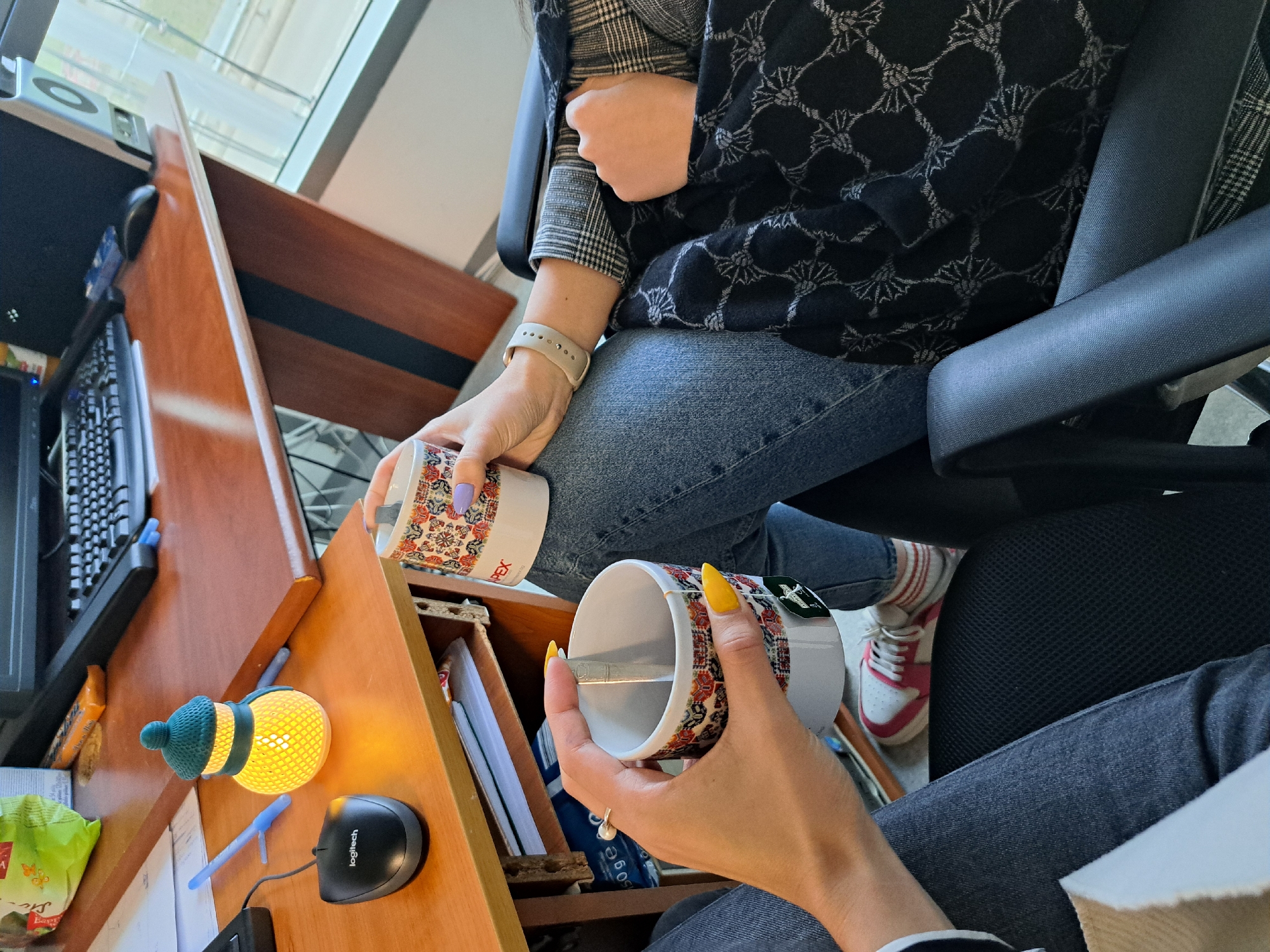 Two individuals are seated at a desk. One person, wearing jeans, a grey-and-white plaid blazer, and a black shawl with a floral pattern, holds a mug with a floral design and purple nail polish. The other, wearing jeans and white and red sneakers, holds an identical mug with floral design and yellow nail polish. The desk has a computer keyboard, mouse, a small nightlight, and various papers and office supplies. The location is likely an office setting in the city of Sofia, Bulgaria.