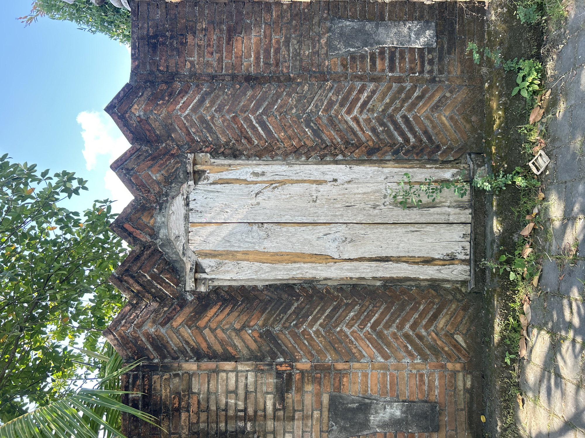 The image shows a weathered, rustic wooden double door set within a decorative brick wall. The wall is constructed with bricks laid in a herringbone pattern on either side of the door, creating a chevron effect that tapers upwards to jagged peaks along the top. The bricks themselves show signs of age, with some discoloration and moss growth. The wooden door planks are vertical and appear to be old and weathered, with visible grain and some chipping. A small amount of green foliage is growing at the base of the door and along the wall. In the foreground, a stone pathway is visible, with scattered leaves and more greenery. Above the wall, the branches of a leafy tree and a blue sky with scattered clouds are seen. The overall impression is one of an aged, possibly historic, entrance in an outdoor setting. This scene is located in Mengwi, Indonesia.