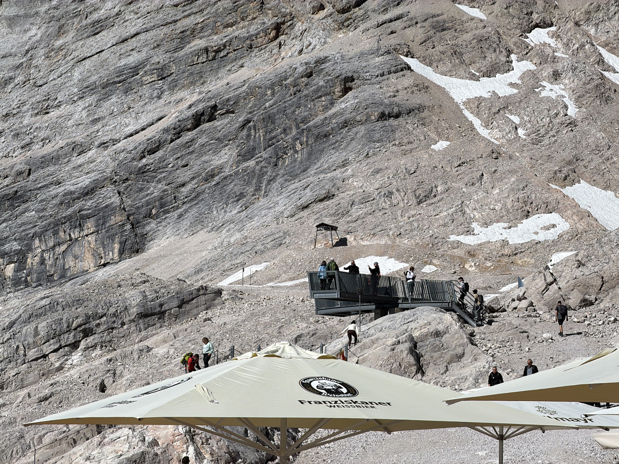 The image depicts a mountainous landscape with scattered snow patches on a rocky incline. In the foreground, two large beige umbrellas with logos are partially visible. A group of people are visible in the midground standing on a modern viewing platform with railings, while others are navigating a rocky path and stairs leading up to it. Further up the mountain, there is a dark structure that appears to be a small building or shelter. The overall environment suggests a high-altitude tourist destination. This scene is likely located at the Zugspitze, Germany's highest peak, which is accessible from Garmisch-Partenkirchen.